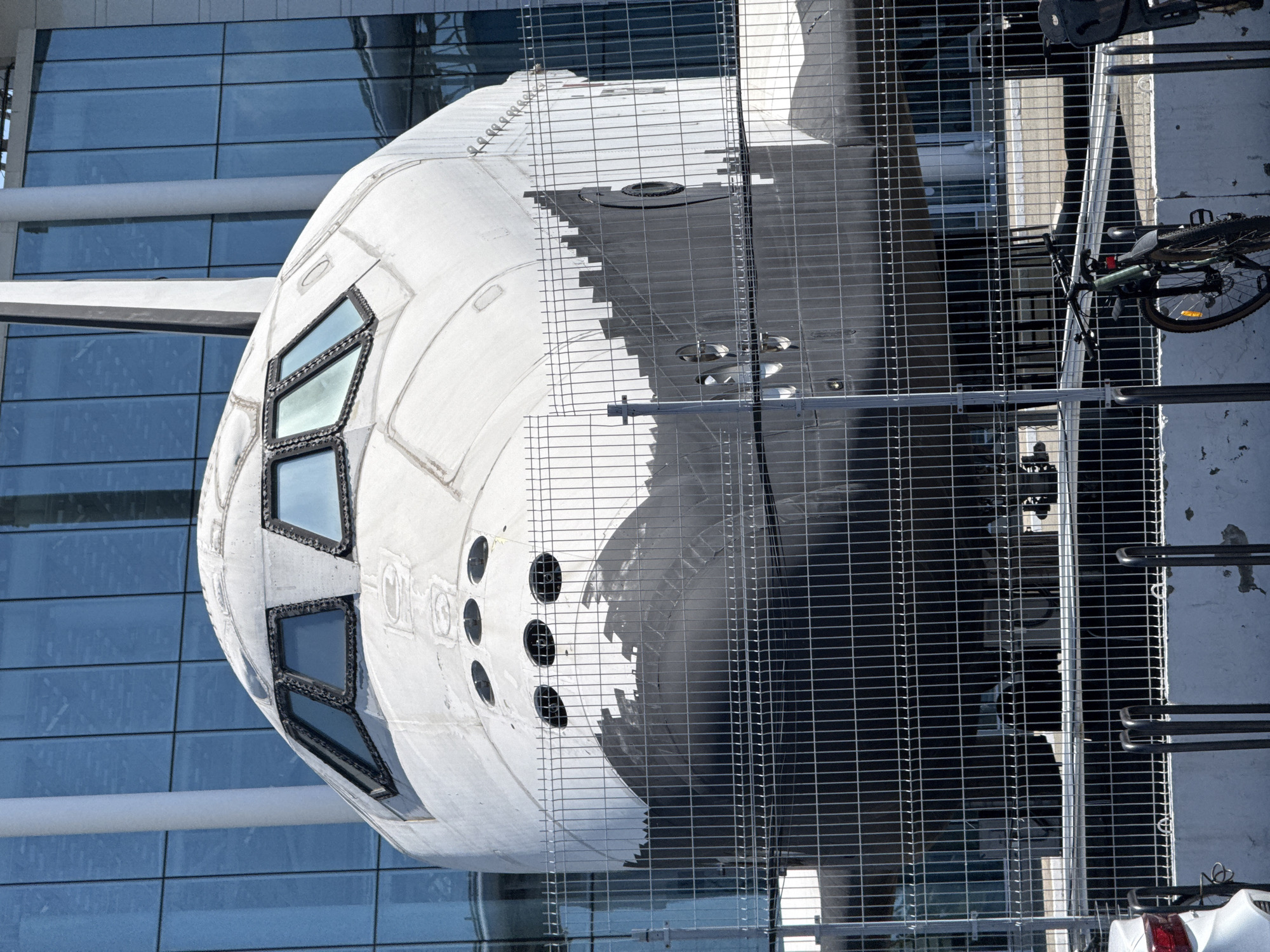 A white, spacecraft-like structure, resembling a space shuttle or a crew capsule, dominates the scene. It features multiple rectangular windows on its side, indicative of a cockpit or observation deck, along with several smaller circular openings further along its fuselage. The surface of the spacecraft appears textured, possibly showing wear or detailed paneling.
In the background, a modern building with numerous reflective blue glass panels creates a striking contrast. These panels suggest a contemporary architectural style, likely housing a museum, science center, or educational institution.
Much of the foreground and midground is covered by a dense metal fence or grating, through which the spacecraft and building are viewed. This barrier partially obscures the view, creating a layered effect. On the far right, behind this fence, a bicycle is partially visible, indicating an outdoor setting where people might be present.
The scene is brightly lit by strong daylight, casting clear shadows, suggesting a sunny day. No discernible activity is taking place; the spacecraft appears to be a static display or exhibit. There is no visible text. Given the location context of Sirius, Russia, this structure likely forms part of a science, technology, or educational exhibition, perhaps related to space exploration.