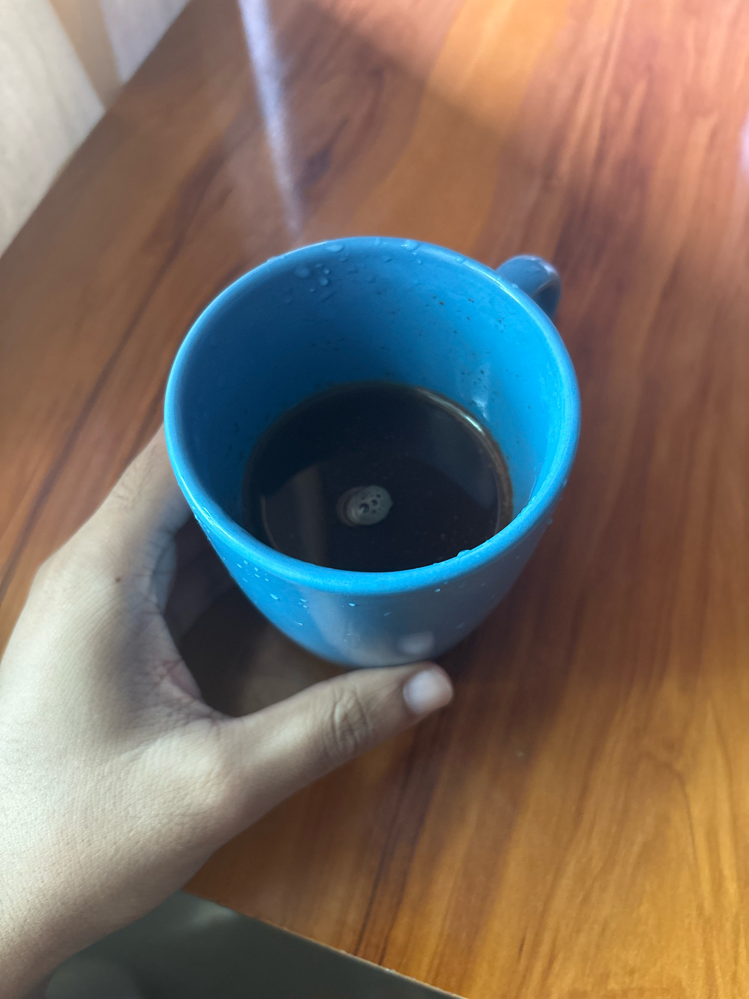 A hand holds a bright blue ceramic mug filled with dark coffee. The mug has condensation on its exterior, indicating it may be cold or freshly filled. The coffee surface shows a small, foamy bubble. The mug rests on a polished wooden surface, likely a table, which exhibits a warm, reddish-brown hue with visible wood grain. The lighting suggests an indoor setting with natural light casting shadows and highlights on the objects. The background is softly blurred, drawing focus to the mug and the hand. The scene is devoid of any specific urban context and thus cannot be localized to Khalagon, India or any other city.