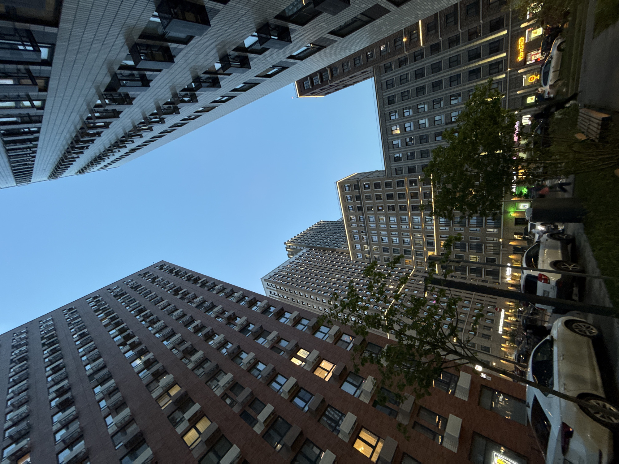The image shows a low-angle view of buildings in Moscow, Russia. The buildings are tall and modern, with multiple windows and balconies. The sky is blue and clear. There are trees and parked cars along the street. People are walking on the sidewalk. Storefronts are lit up.