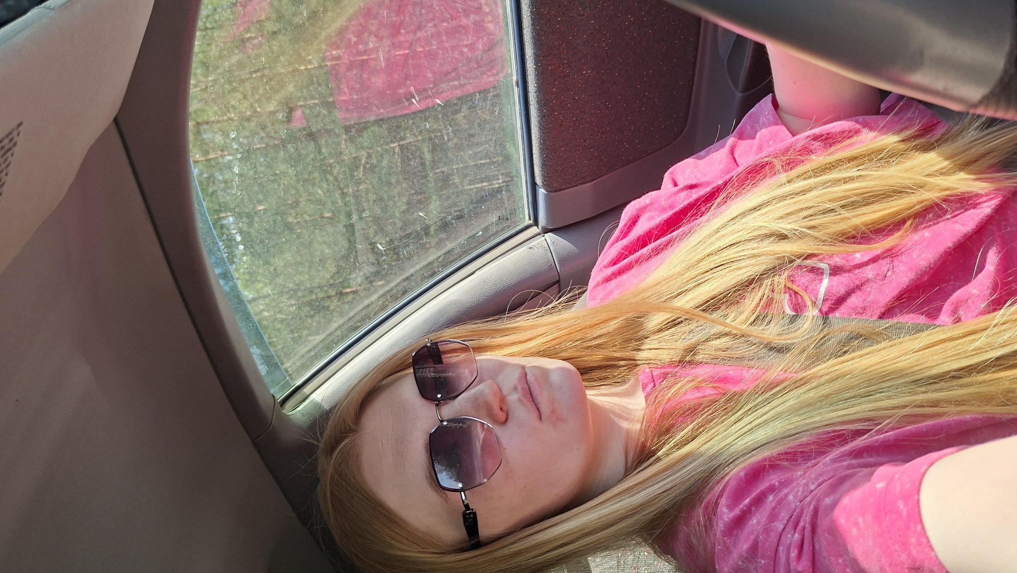 The image captures a light-skinned person with blonde hair wearing sunglasses and a pink shirt, seemingly lying down inside a vehicle. The location appears to be inside a car, judging by the window, interior panels, and a portion of the steering wheel visible. The exterior view through the window is blurred, suggesting movement. The scene does not provide sufficient information to confirm that the location is Chaikovsky, Russia.