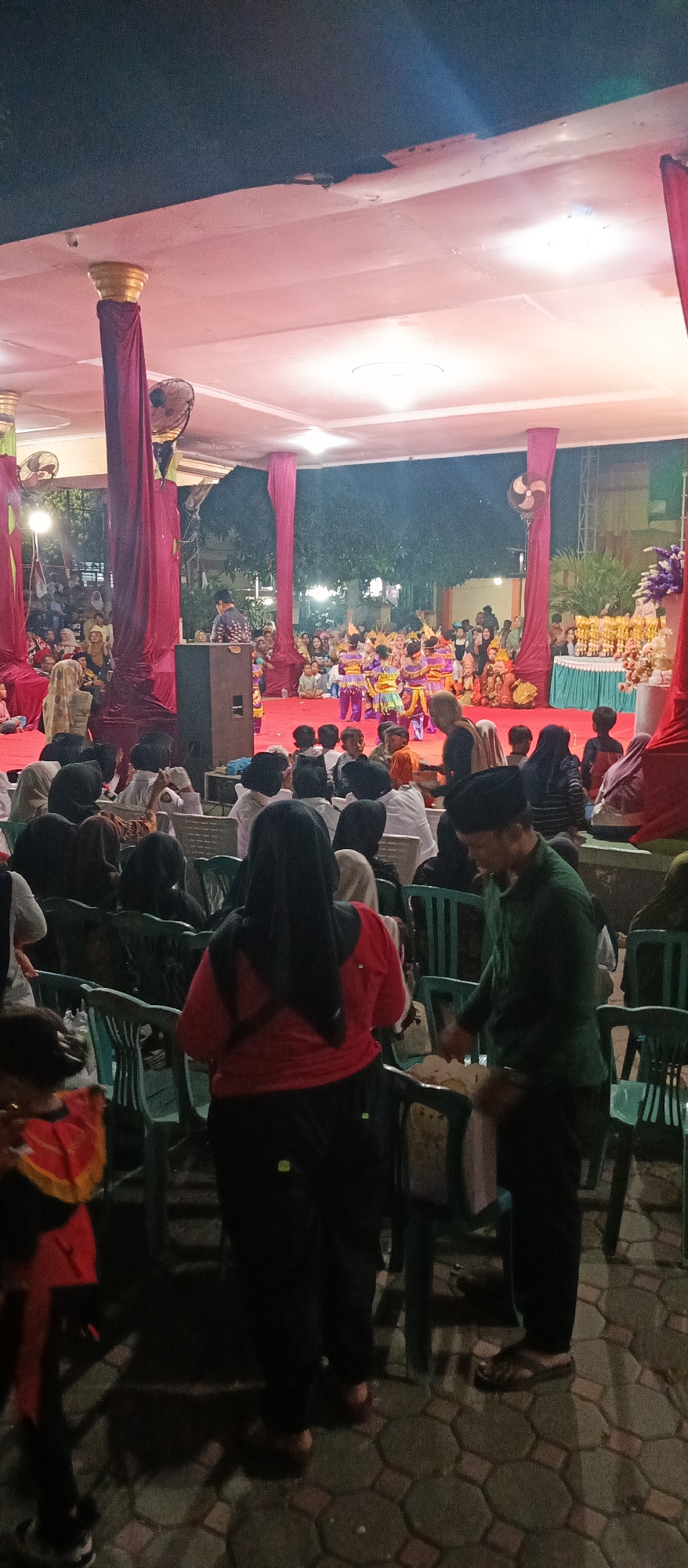 An outdoor evening event is underway in Arjasa, Indonesia, featuring a performance on a stage adorned with red drapes and illuminated by warm lights. A group of children in colorful traditional attire are actively engaged in a dance, with several adults in the audience observing from seated positions on plastic chairs arranged on a paved ground. A man in a green shirt is interacting with individuals seated in the audience. The backdrop of the stage is partially visible, showing more spectators and decorative elements, possibly trophies, arranged on a table. The lighting creates a slightly hazy atmosphere, typical of an evening outdoor gathering.