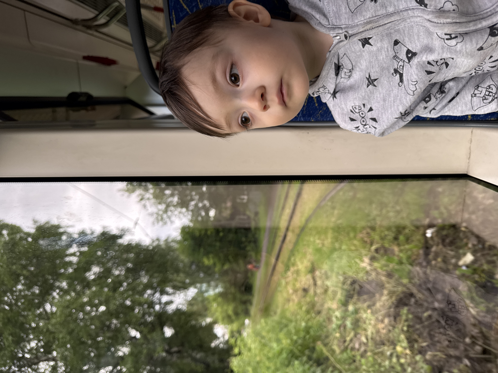 A young child with brown hair and brown eyes looks out a window from inside a vehicle. The child is wearing a gray zippered hoodie with a cartoon print. Outside the window, there are green trees and grass, and train tracks. In the distance on the tracks, a person in an orange shirt is visible. The scene appears to be on a tram in Izhevsk, Russia.