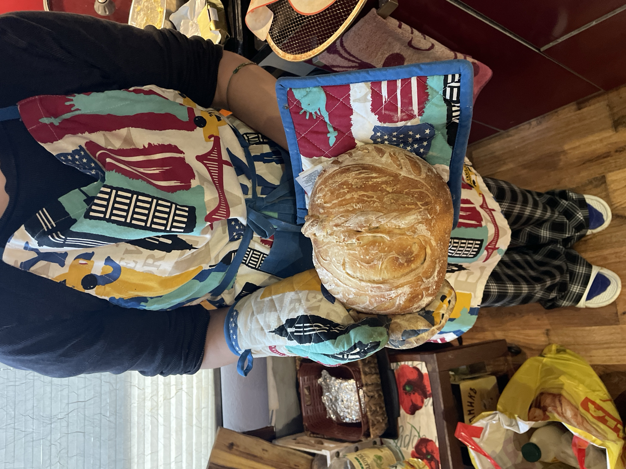 The image shows a person holding a round loaf of bread with both hands, protected by oven mitts and a pot holder. The person is wearing a dark long-sleeved shirt and an apron with a colorful, abstract pattern resembling elements of the American flag. They are also wearing plaid pants and slippers. The background is a cluttered kitchen setting with wooden floors. A bag is on the floor and a shelf with canned and boxed goods is visible. The location is not discernable from the image and the suggestion that the scene is in Burgas, Bulgaria, cannot be confirmed from the provided visual data.