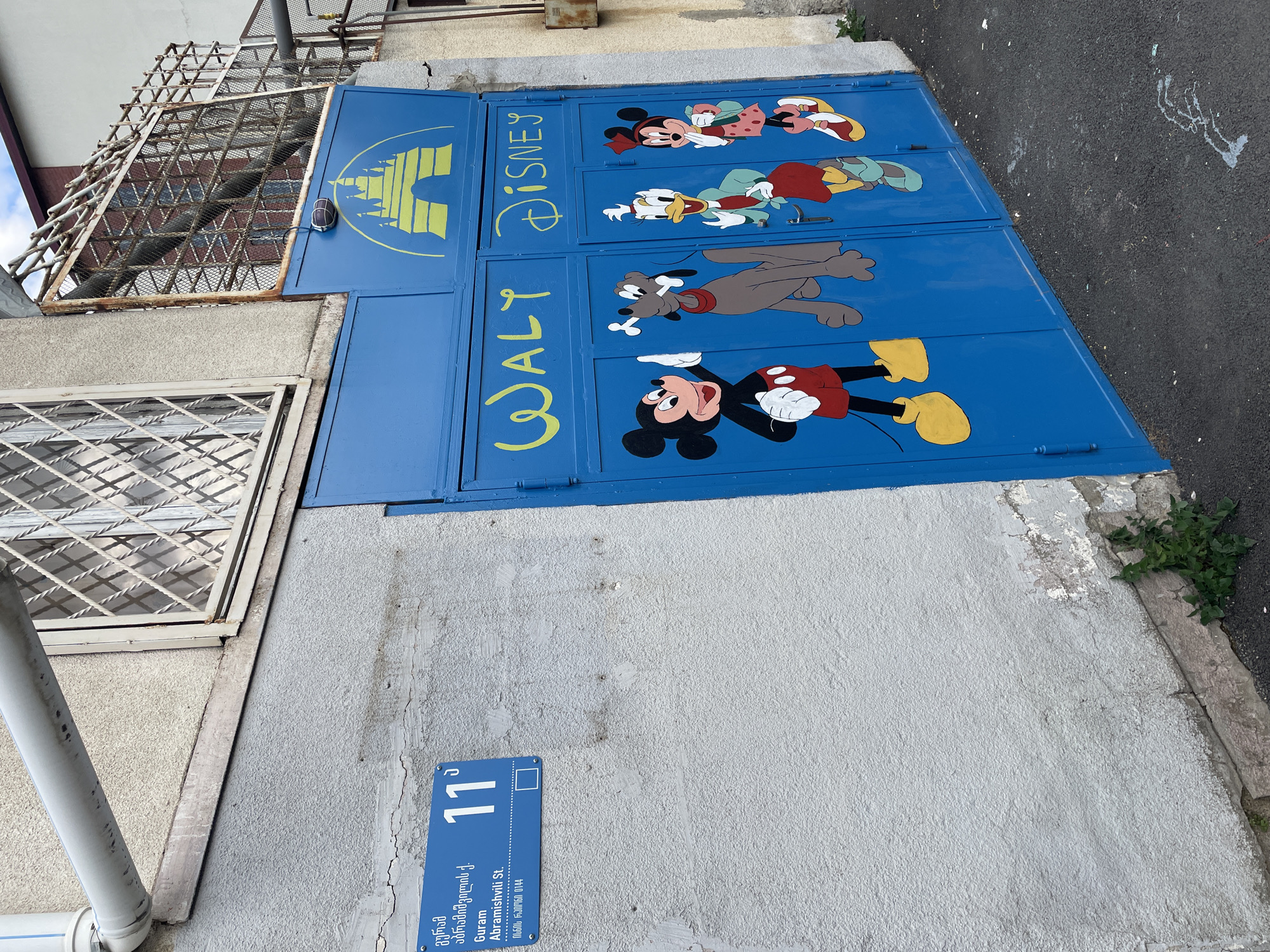 The image shows a building in Tbilisi, Georgia, at the address Guram Abramishvili Street 11. The building's gray exterior wall has a blue street number sign and a blue metal gate adorned with artwork of Disney characters Mickey Mouse, Minnie Mouse, Donald Duck, and Goofy. The words "WALT DISNEY" are painted in yellow on the gate. The gate is composed of four panels stacked on top of each other. A window with a white frame and security bars is on the left side of the gate.