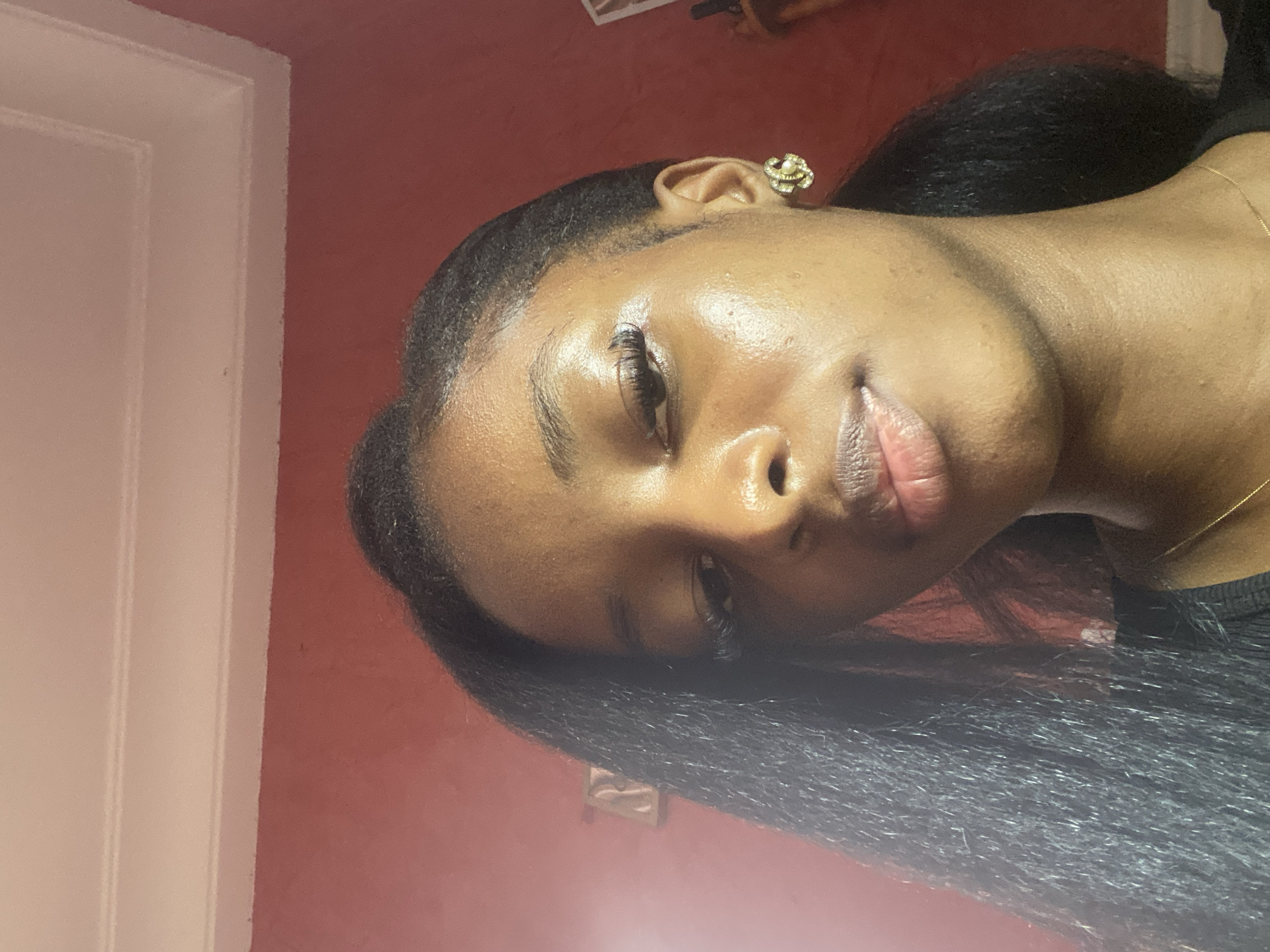 A close-up, overhead shot shows a young Black woman with dark hair and light skin against a red wall with a white ceiling. She is looking down and to her left, her gaze slightly obscured by her hair. She is wearing a gold earring with a pearl center and a thin gold chain necklace. The woman's skin is smooth and appears to have a subtle sheen. Her hair is long and dark, styled in a way that parts on the left and falls over her shoulder. The background is a plain red wall with white molding at the top, suggesting an indoor setting. The overall lighting is soft, with a noticeable highlight on her forehead and cheekbone. There are no discernible city landmarks or indications of Jos, Nigeria, in the image.