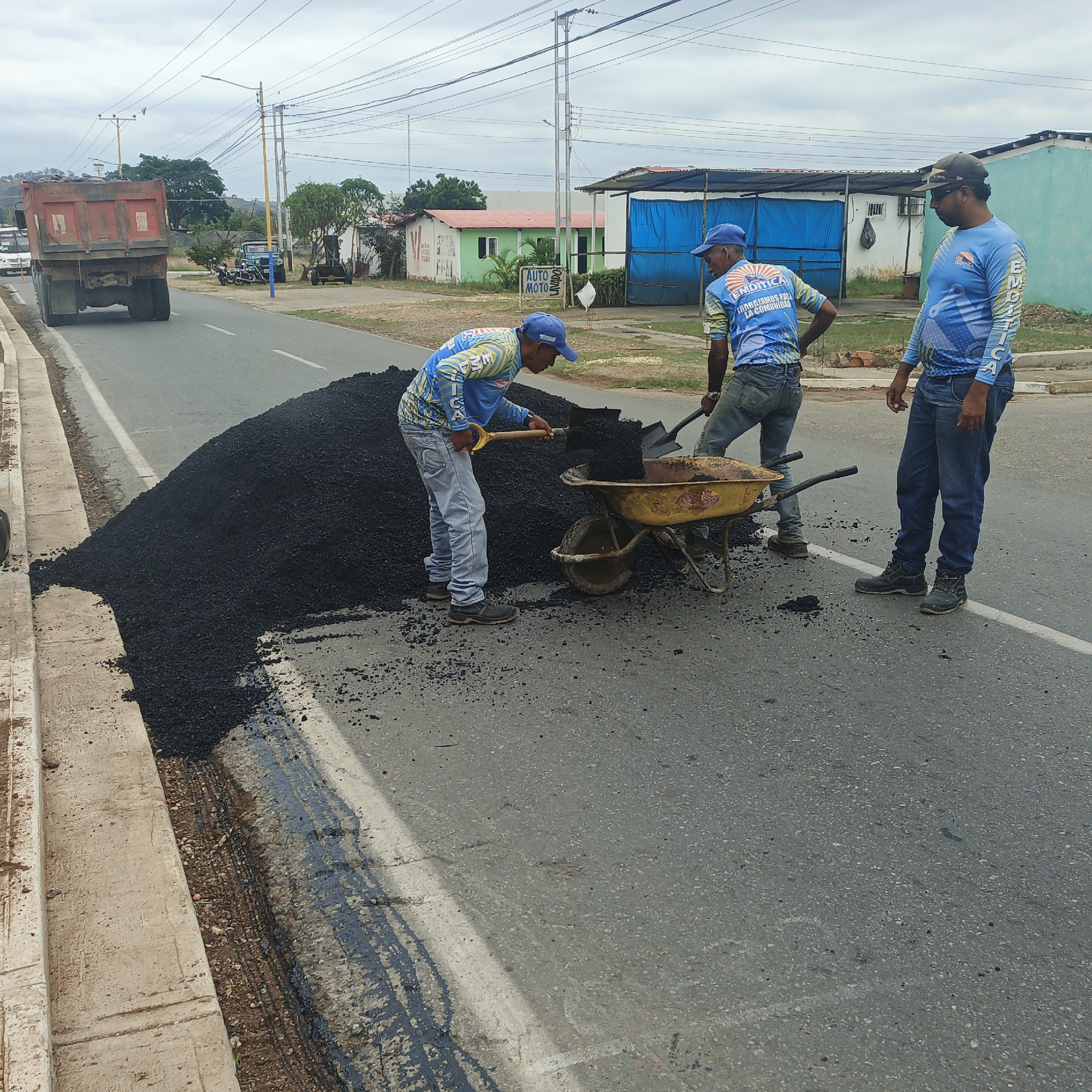 Three men are repairing a road in Tinaquillo, Venezuela. One man is shoveling asphalt from a large pile into a yellow wheelbarrow. Another man is also shoveling asphalt. The third man stands beside them, watching. All three men are wearing similar blue shirts with an emblem. There is a red dump truck parked on the side of the road nearby. The background includes buildings, power lines, and other vehicles on the road. The sky is overcast.