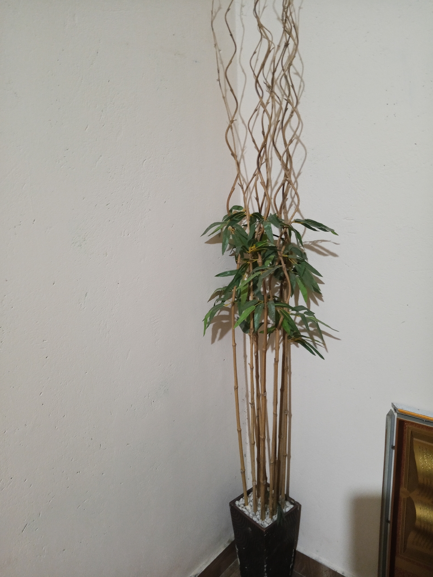 A tall, decorative faux bamboo plant stands in a dark, square planter filled with white pebbles. The plant consists of numerous thin, brown stalks that twist upwards, some with scattered green leaves. The planter is positioned in a corner against a textured, off-white wall. To the right of the planter, a portion of a decorative object with a golden pattern and a dark brown frame is visible. The floor appears to be tiled with a wood-grain pattern. The image was taken indoors, likely in a residential or commercial space in Lubuklinggau, Indonesia.