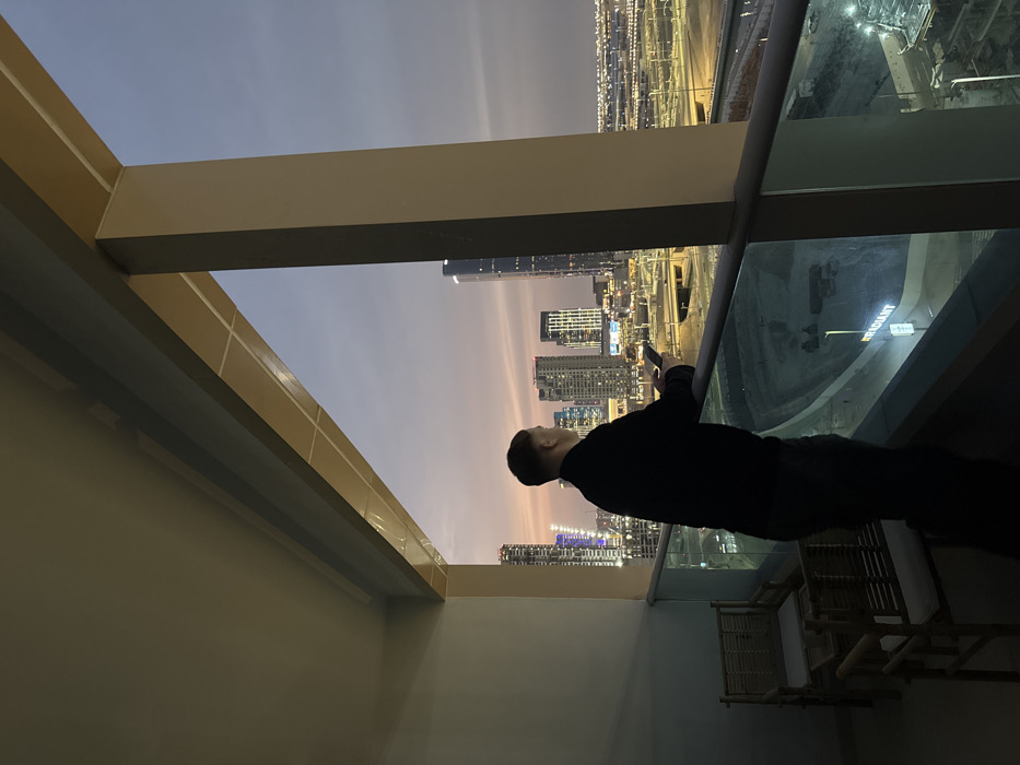 The image depicts a high-altitude view from a balcony on Al Reem Island, Abu Dhabi, United Arab Emirates, at twilight.
A single adult male, viewed from the back, stands at the balcony railing, looking out over the city. He wears dark clothing and holds a smartphone in his right hand, with his forearms resting on the glass-paneled railing. To the immediate left of the subject, two small, light-colored chairs with white cushions and a matching small table are visible on the balcony.
Beyond the balcony, a vast urban landscape unfolds under a sky transitioning from orange and pink hues near the horizon to a darker blue overhead. Numerous high-rise buildings, many illuminated, populate the middle ground and distance. Below the balcony, a significant construction site is visible, featuring excavated ground and construction elements. A prominent white sign bearing the partially visible word "RADIANT" is situated within this construction area. A complex network of roads and overpasses can be seen traversing the cityscape. Artificial lights from buildings, streets, and the construction site are active, indicating either dusk or dawn.