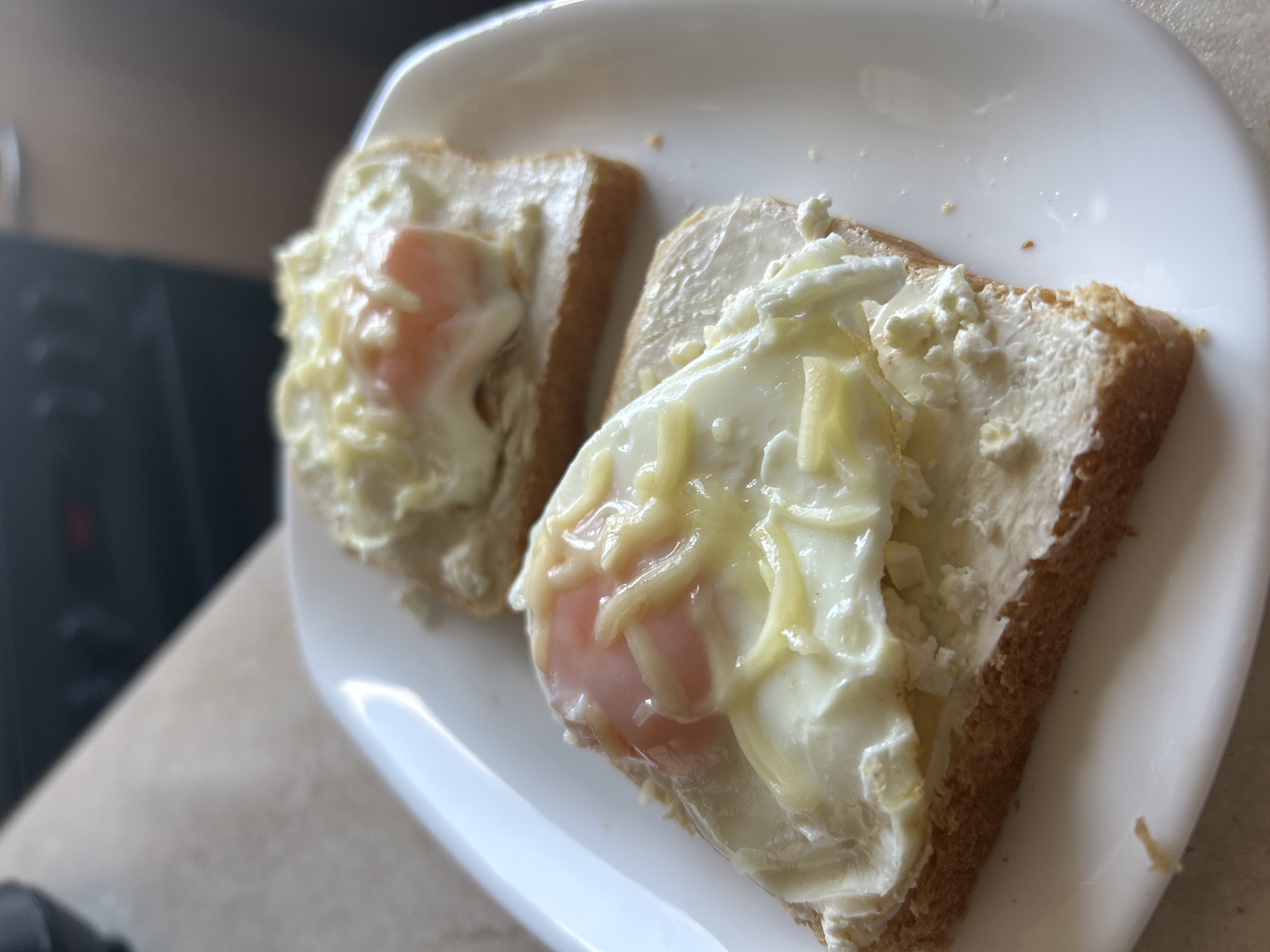 Two slices of toasted bread, each generously spread with a white, creamy substance and topped with a single fried egg, are presented on a glossy white, rectangular ceramic plate. The egg yolks exhibit a soft, pinkish-orange appearance, indicative of a runny or soft-boiled state, and are covered with a layer of shredded, partially melted light-yellow cheese. The plate rests on a light-colored, subtly textured countertop, consistent with a kitchen environment. In the blurred background, dark kitchen appliances or cabinetry are visible, reflecting ambient light. No individuals are present. The scene is located in Sofia, Bulgaria.