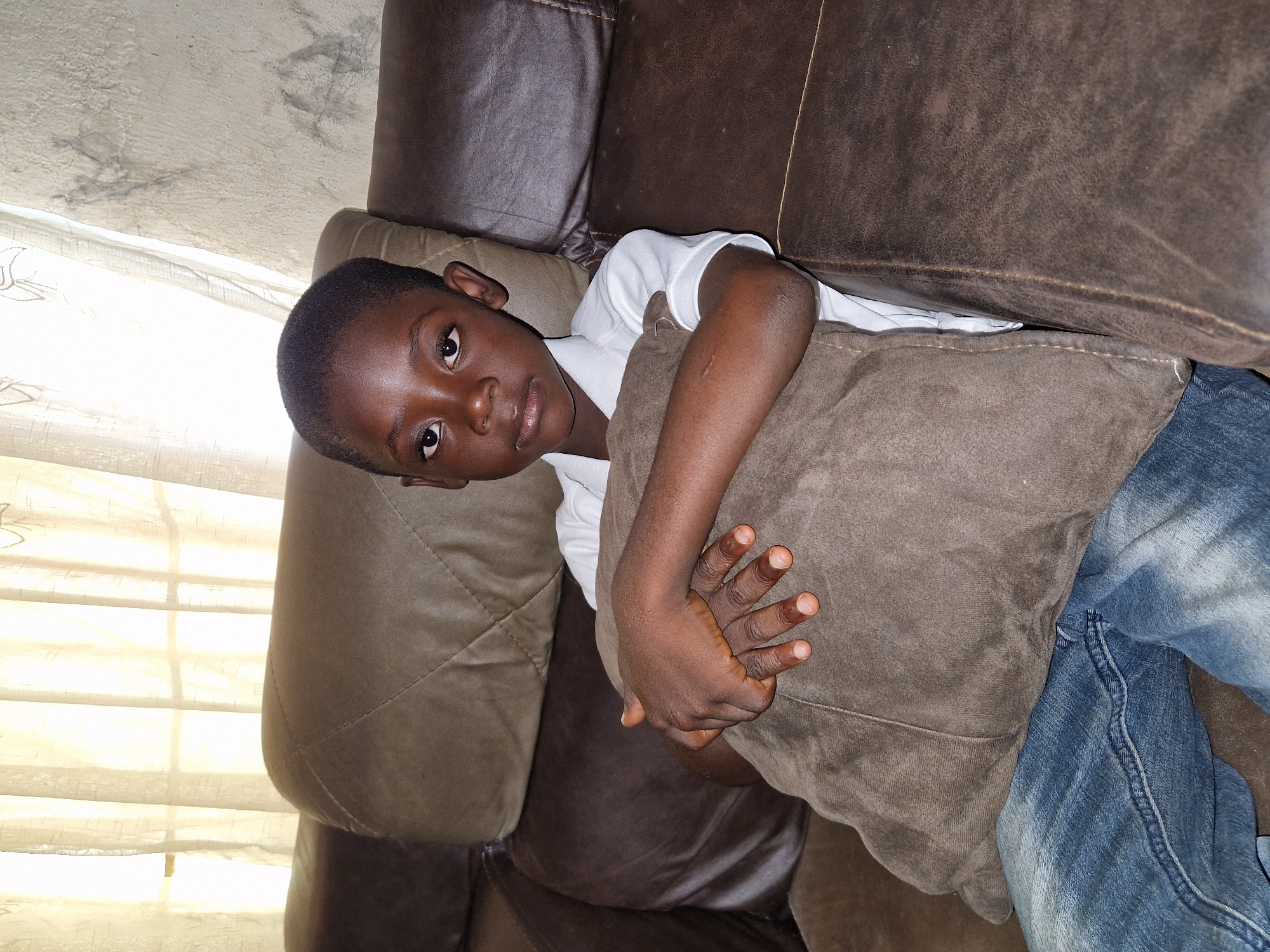 A young Black boy is seated on a brown leather sofa, embracing a brown pillow. He is wearing a white collared shirt and blue denim jeans. The boy is positioned in the center of the frame, with the sofa extending to the left and right. Behind him, a light-colored curtain is visible, with a wall on the right side of the frame. The lighting suggests an indoor setting, likely a living room. The scene appears to be in Jos, Nigeria, given the context provided.
