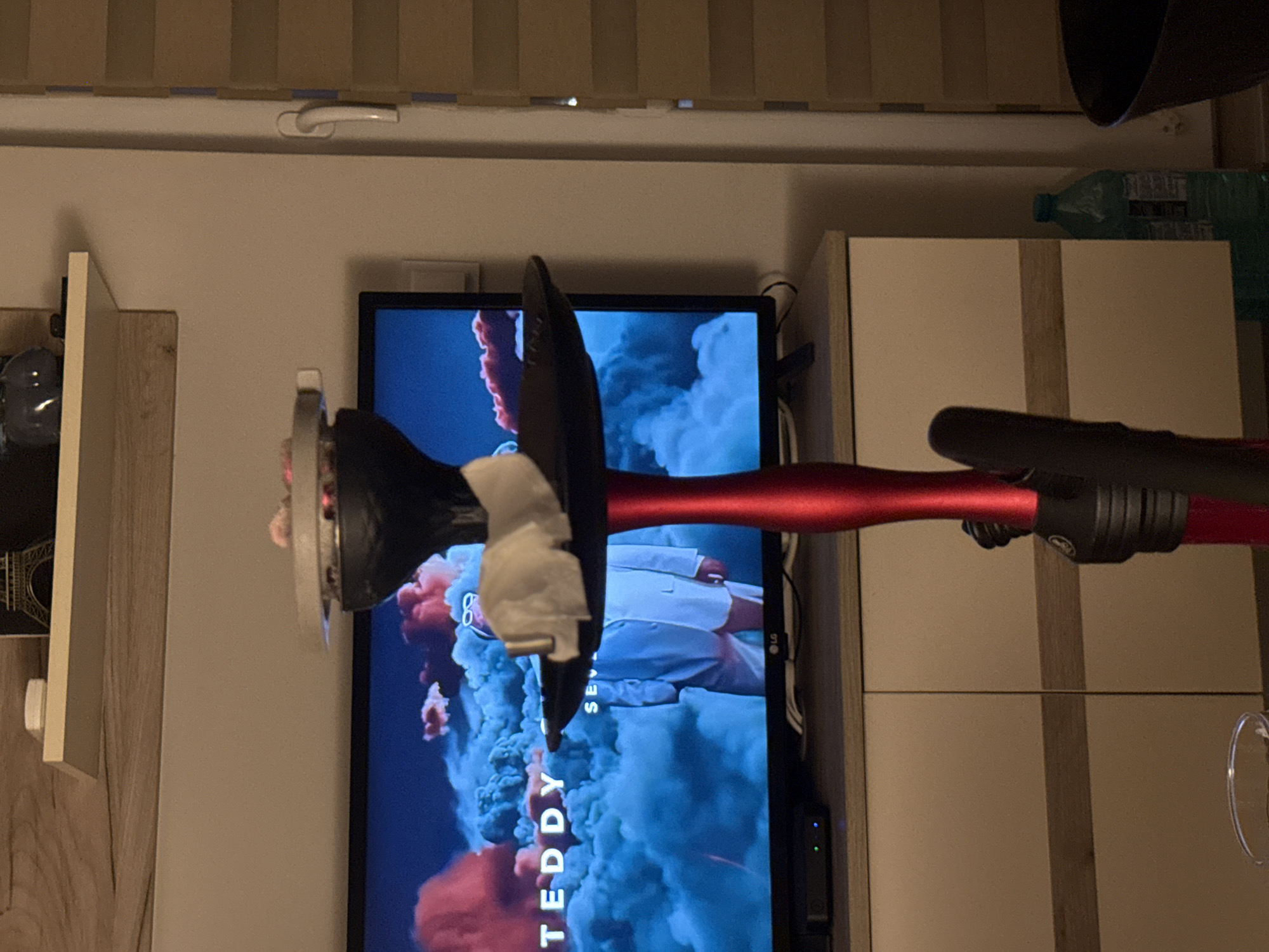 This is a medium shot, eye-level photograph of a red hookah standing in front of a television screen. The TV is displaying a scene with clouds and the text "TEDDY" and "SEV." The hookah has a black bowl on top, with what appears to be white paper or cloth covering the coals. The background consists of a wooden shelf above the TV, a white cabinet below, and a vertical blind on the right side of the frame. A green bottle of water and a black container are visible on the floor to the right of the cabinet. The image appears to have been taken indoors, in a domestic setting, possibly a living room. There is no indication of the specific location in Varna, Bulgaria.