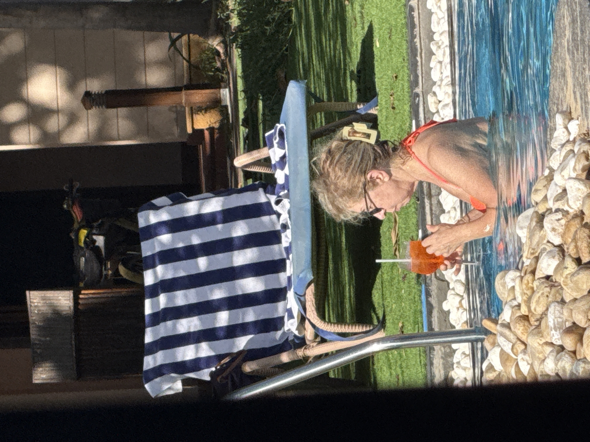 An elderly Caucasian woman with blonde hair and glasses is in a swimming pool, holding an orange cocktail with a straw. She is wearing a red bikini top. The pool is surrounded by large white and tan rocks. In the background, there is a lounge chair with a blue and white striped towel draped over it, a grassy area, and a building with wooden doors and potted plants. The scene appears to be outdoors, possibly at a resort or hotel, given the lounge chair and swimming pool. The lighting suggests it is daytime, with sunlight casting shadows. The presence of a lounge chair and poolside setting is consistent with a vacation or leisure environment, often found in Dubai, United Arab Emirates, a city known for its luxury hotels and resorts.