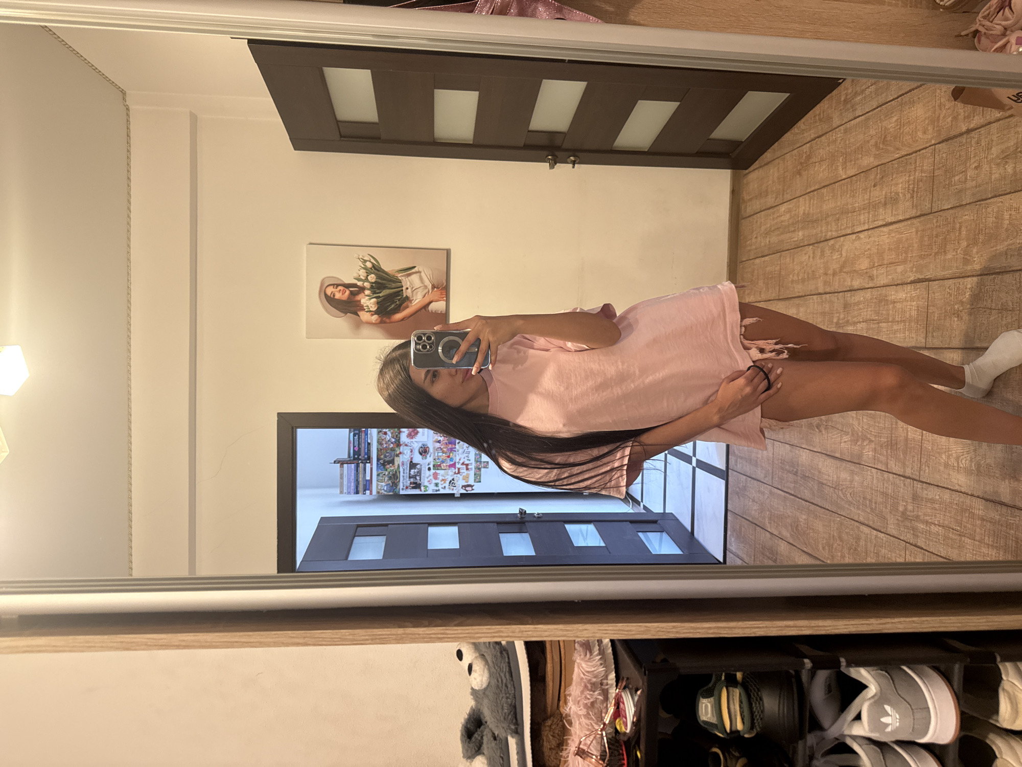 The image shows a young woman taking a selfie in a mirror. She is wearing an oversized pink t-shirt and white socks. Her long, dark hair is down. She is standing in what appears to be a hallway with wooden flooring. To her left, there is a shoe rack with several pairs of shoes and some plush slippers. A large mirror is positioned in front of her, reflecting the scene. On the wall behind her, there is a framed picture of a woman holding tulips. To her right, there is a dark wooden door with frosted glass panels. The environment suggests a residential setting, possibly an apartment. The image was taken in Chișinău, Moldova.