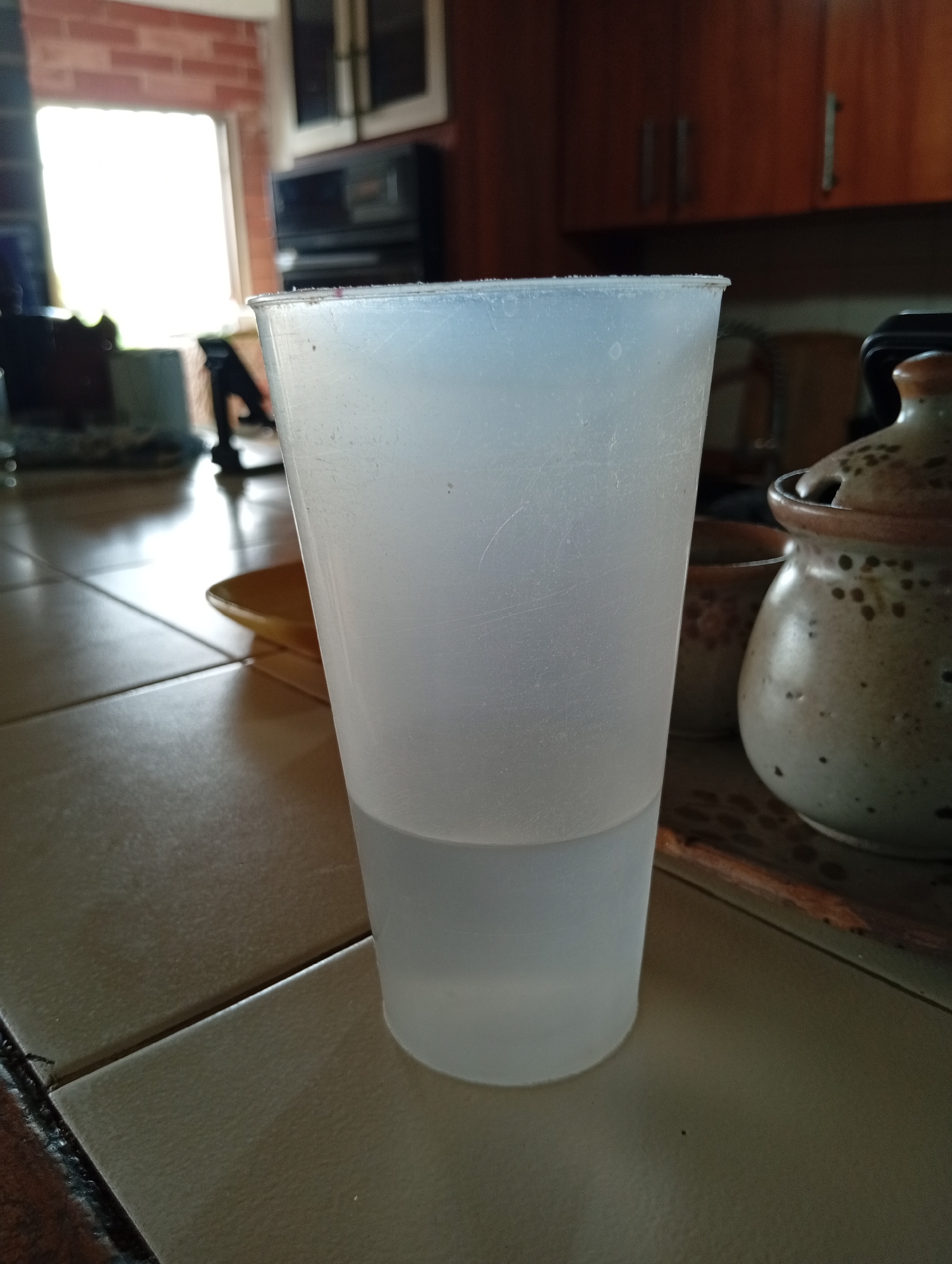 A close-up, eye-level, slightly angled shot captures a translucent plastic cup filled with clear liquid to approximately half its volume, resting on a tiled kitchen counter. The cup, positioned in the foreground and slightly off-center to the left, exhibits some visible scratches. Behind the cup, to the right, a ceramic pot with a lid and a speckled pattern sits on a wooden surface, with another similar, smaller pot visible behind it. The background reveals a kitchen environment with wooden cabinets and a window allowing bright natural light to enter, illuminating the scene and casting reflections on the tiled floor. The overall setting suggests an interior of a residential kitchen in San Antonio de Los Altos, Venezuela.