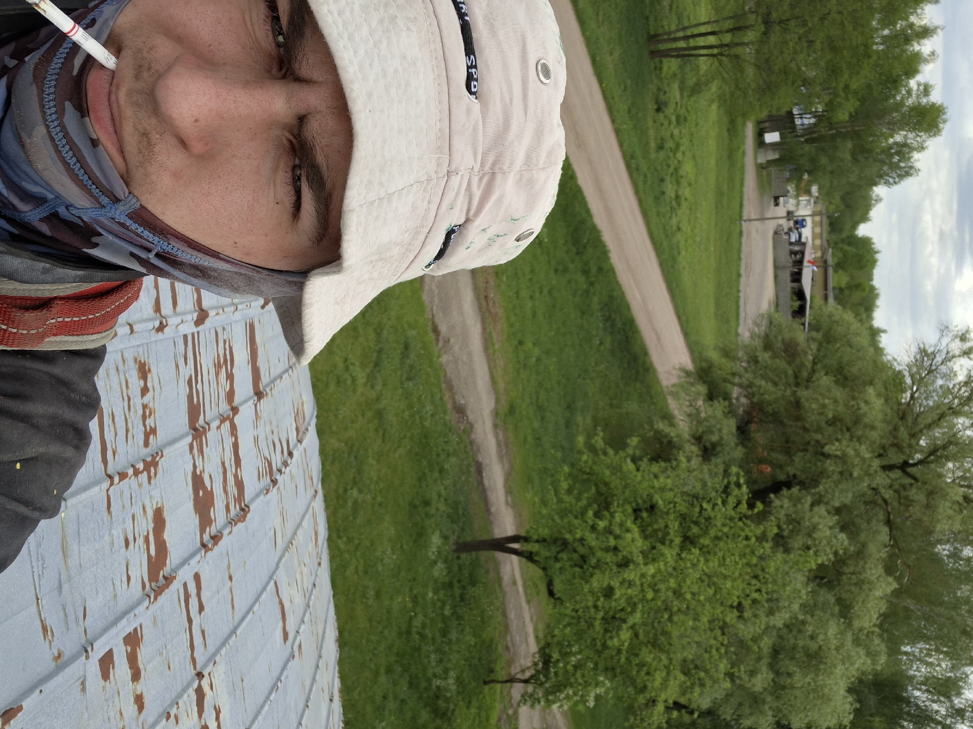 A selfie of a man wearing a white hat, a camouflage balaclava, and what appears to be work gear, potentially a harness, is taken from an elevated position, possibly on a metal roof with rust. The background shows a grassy area with trees and a dirt road leading to a building complex in the distance. The city of Zhabinka, Belarus, is not readily discernible from the image's visual content.