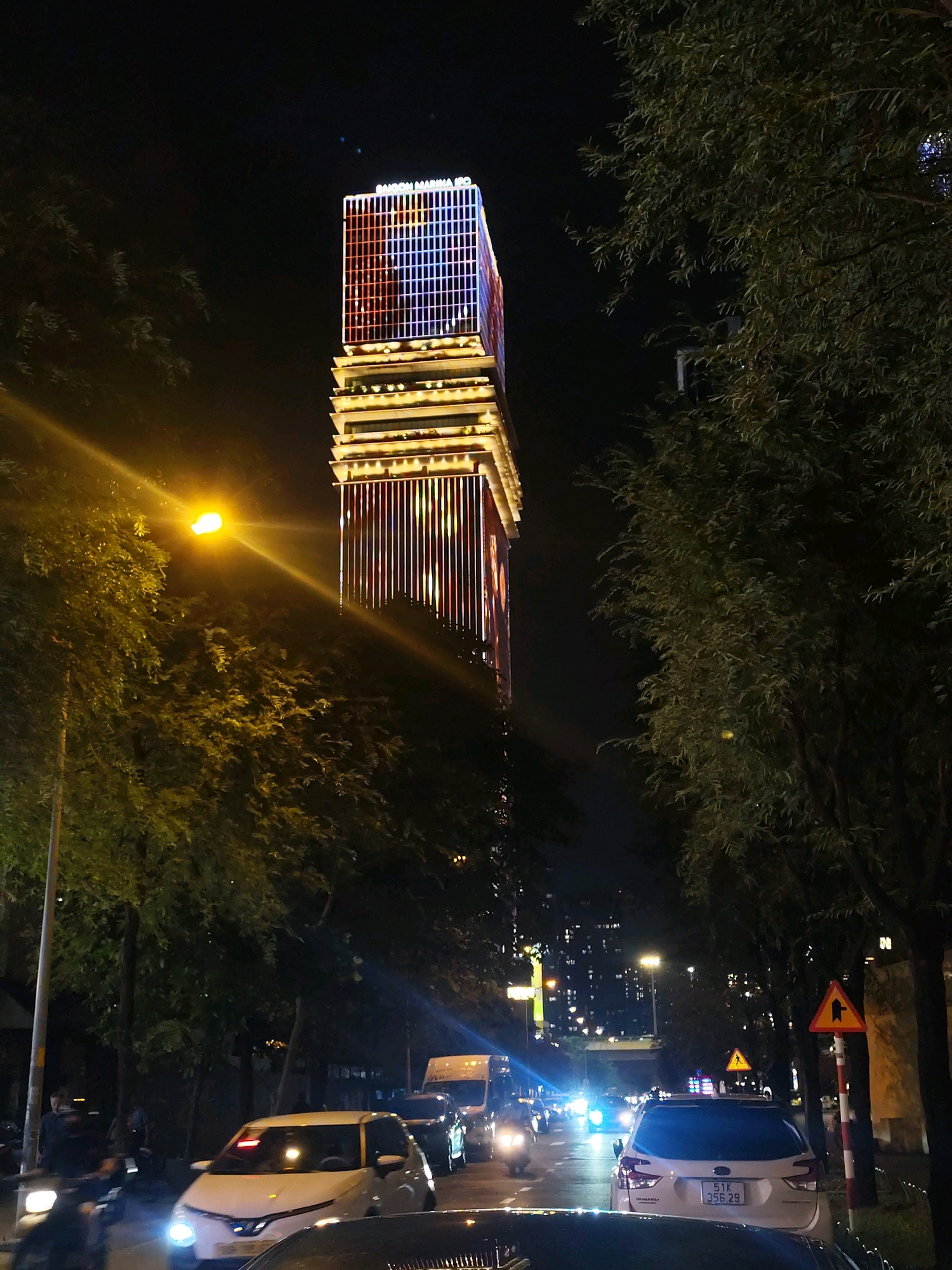 This is a night-time street-level photograph taken in Ho Chi Minh City, Vietnam, likely in the vicinity of Saigon Marina IPO, a tall, illuminated skyscraper visible in the background. The building is adorned with red and blue lights on its upper section and vertical yellow and red lights on its lower sections, creating a striking visual effect against the dark sky. The street is lined with mature trees, their foliage illuminated by streetlights and the building's lights, casting dappled shadows. A bright streetlamp on the left side of the frame emits a strong yellow glow. Several vehicles are present on the road, including cars and motorcycles, with their headlights on, indicating traffic. One white car in the foreground has its headlights on, and a red illuminated sign is visible on its windshield. Another white car further down the street has a Vietnamese license plate reading "51K 356.29". Two traffic signs are visible on the right side of the road: a yellow triangular sign with a black arrow indicating a right turn and a straight path, and another similar sign further back. The scene depicts a busy urban environment at night, characterized by artificial lighting from buildings and vehicles, with a prominent architectural landmark dominating the skyline.