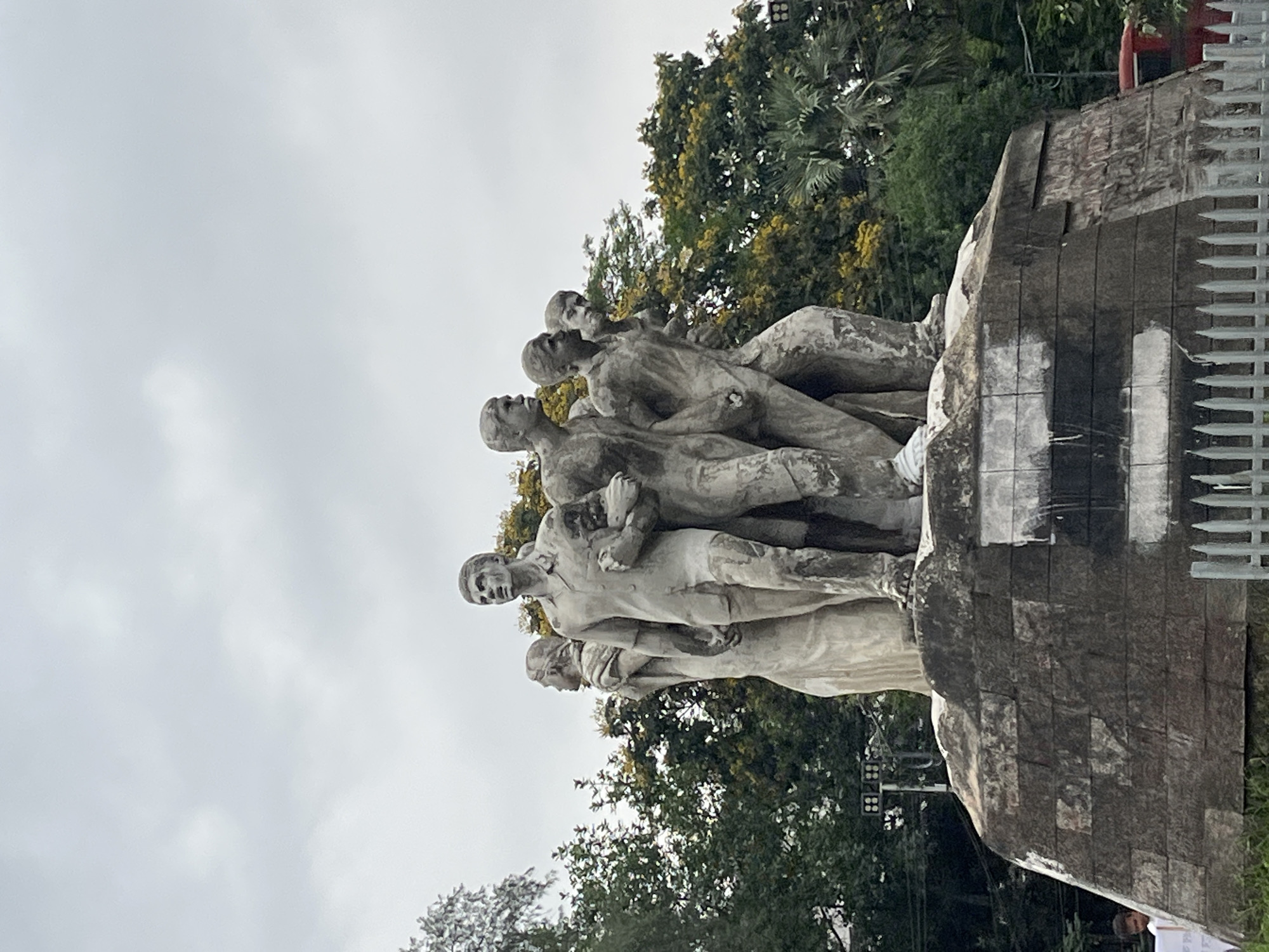 A large, weathered concrete sculpture, "Aparajeyo Bangla" (Invincible Bengal), is depicted under an overcast sky in Dhaka, Bangladesh. The monument features seven human figures, predominantly male, rendered in a realistic, dynamic style. The figures appear to be pushing forward, conveying a sense of collective struggle, determination, and forward momentum. Their expressions are resolute, and their bodies are lean, suggesting physical effort or an allegorical representation of resilience.
The sculpture is set upon a robust, rough-hewn concrete base that shows signs of weathering and some dark markings. Lush green and yellowish trees form the background, partially obscuring other structures. The sky is a uniform, light grey, indicating an overcast day. To the right of the sculpture, a modern, light-colored metal fence or barrier is visible. In the bottom right corner, a small, unidentifiable portion of a white sign or banner with text, likely in Bengali, is partially visible, along with a glimpse of a person. The monument's overall appearance suggests it is a significant public artwork commemorating a historical event or movement.