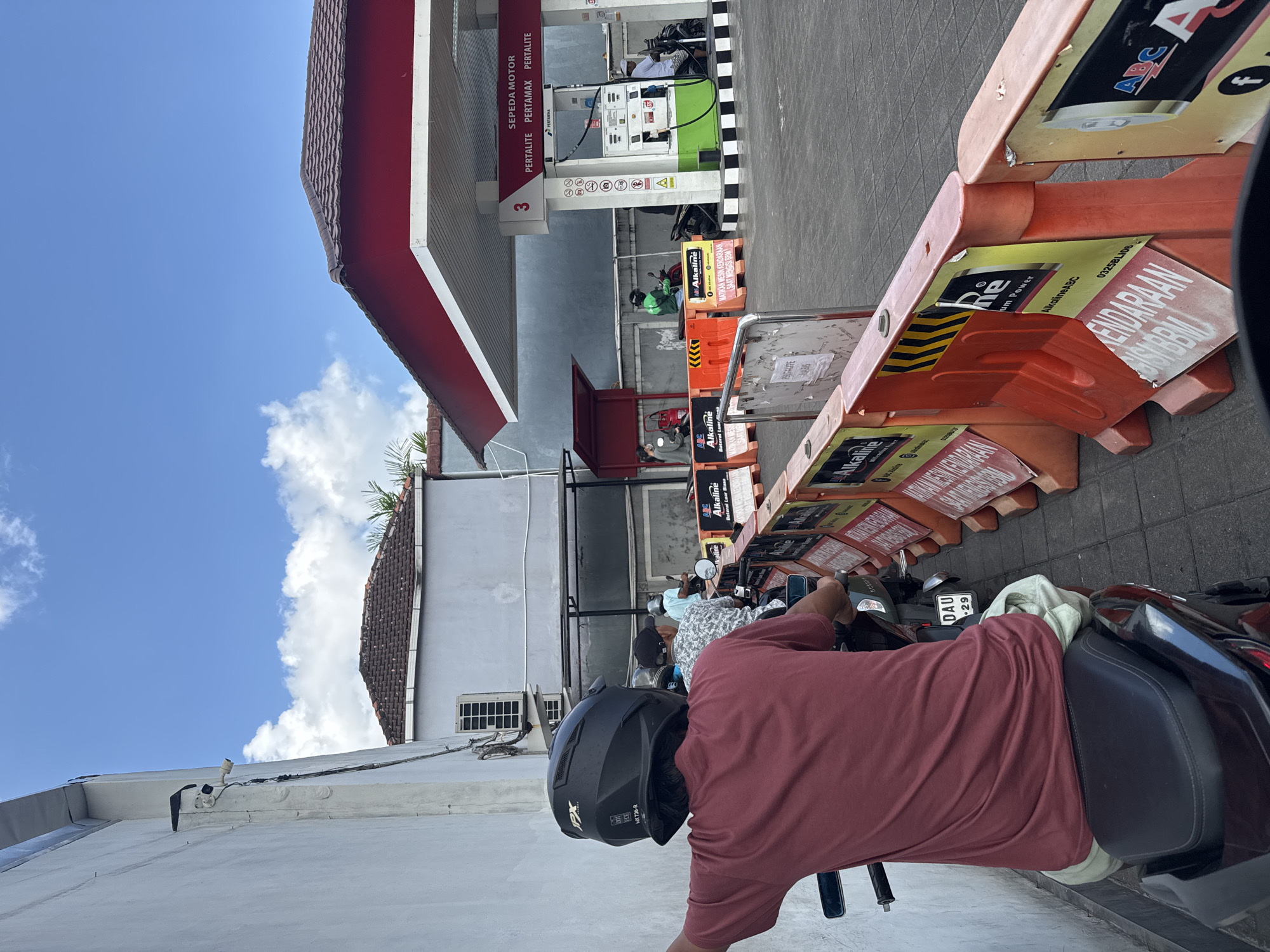 The photograph was taken in North Kuta, Indonesia. It captures a man sitting on a black motorcycle at a Pertamina gas station. He is wearing a red shirt and a black helmet with "IXX MX726-R" printed on the side. His motorcycle's license plate reads "DAU 29." In the background, other people are waiting in line on motorcycles to get gas. The gas station has a red and white awning and gas pumps labeled "PERTALITE PERTAMAX PERTALITE." Orange and yellow traffic barriers with "Alkaline ABC" and "MATIKAN MESIN KENDARAAN SAAT MENGISI BBM" (Turn off the vehicle engine when filling up fuel) signs line the lane leading to the pumps. The sky is blue with some clouds.