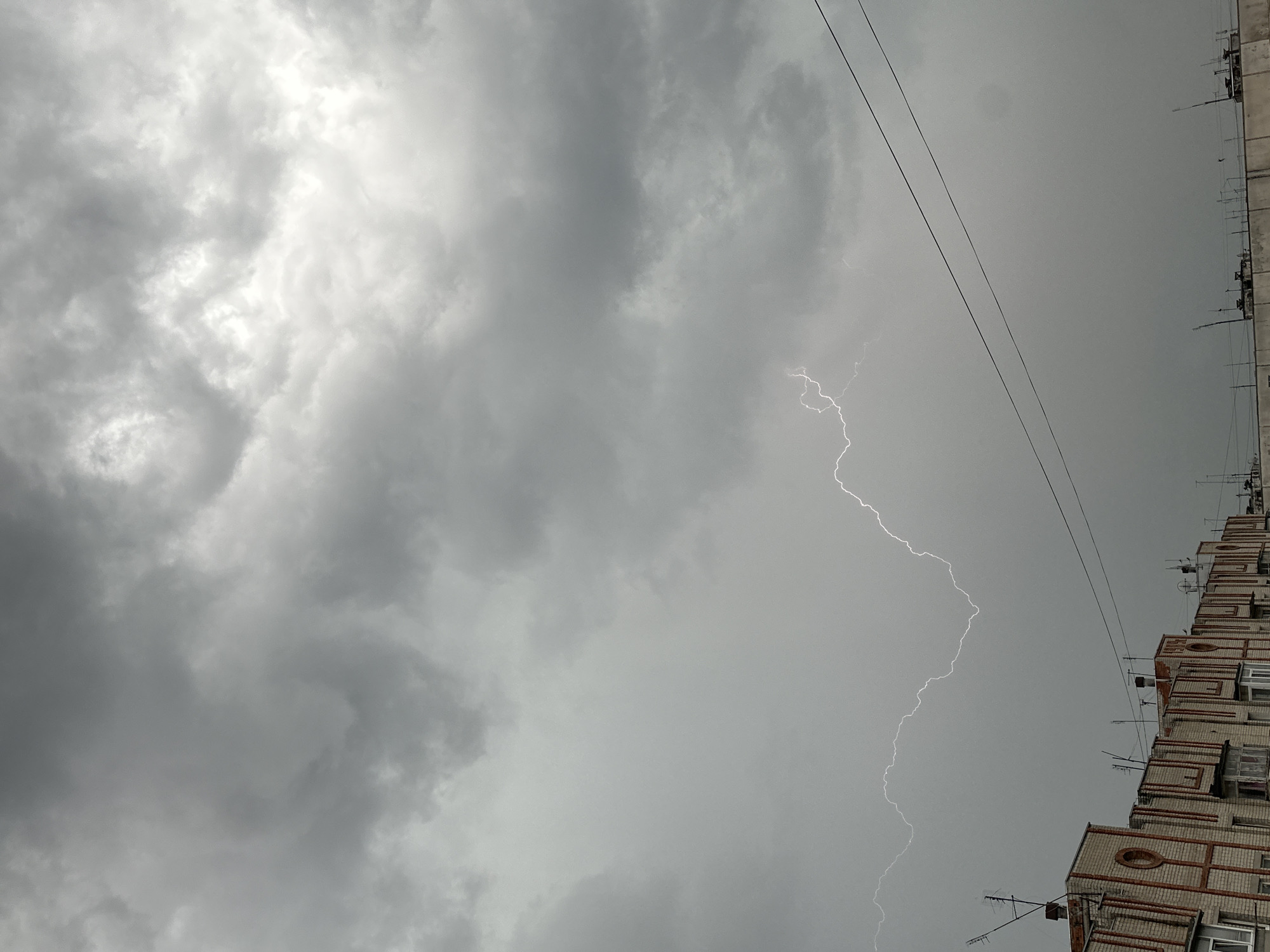 The image depicts a lightning strike during a storm in an urban environment. Dark, heavy storm clouds dominate the sky, obscuring much of the ambient light. A bright bolt of lightning is visible, forking down from the clouds toward the ground. In the lower right corner, a section of a multi-story brick building is visible, including several antennas protruding from the roof and sides of the building. Power lines are also visible in the sky.