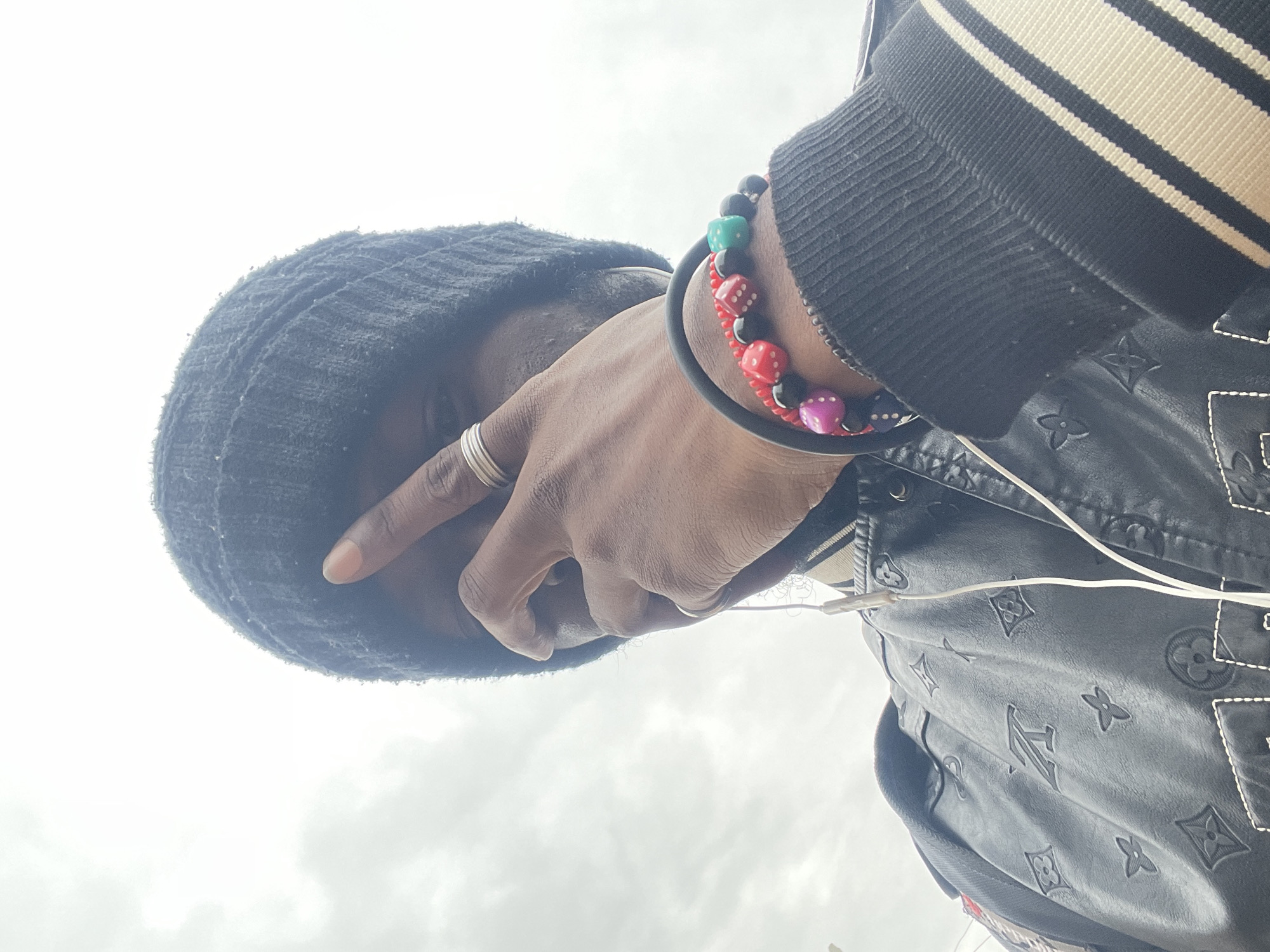 A low-angle, close-up, eye-level medium shot captures a person from the chest up, with the sky serving as a bright, overcast backdrop. The subject is wearing a black beanie and a black jacket with a repeating embossed pattern resembling the Louis Vuitton monogram. The jacket also features striped cuffs on the sleeves. Two silver rings are visible on the subject's right hand, which is positioned in front of their face, obscuring the eye. A multi-colored beaded bracelet with dice-shaped beads is worn on the wrist. White wired earbuds are seen with the wires trailing down. The setting appears to be outdoors, with the cloudy sky suggesting an ambient light condition. No specific landmarks are visible to pinpoint the location within Jos, Nigeria.