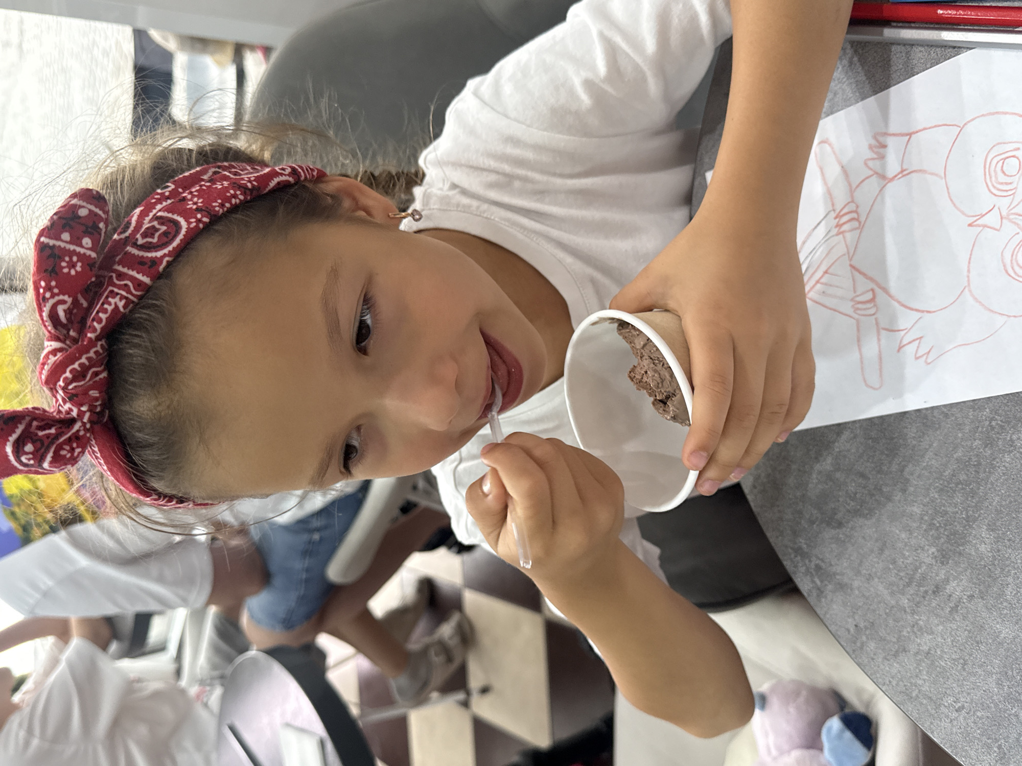 A young girl with a red bandana tied around her head is eating ice cream from a paper cup with a plastic spoon. She is wearing a white t-shirt and a small hoop earring with a pink charm. A drawing of an owl is on the table in front of her. In the background, other people are visible, and the floor has a checkered pattern. The scene appears to be taking place in an indoor establishment, possibly a cafe or restaurant, in Alushta, Ukraine.