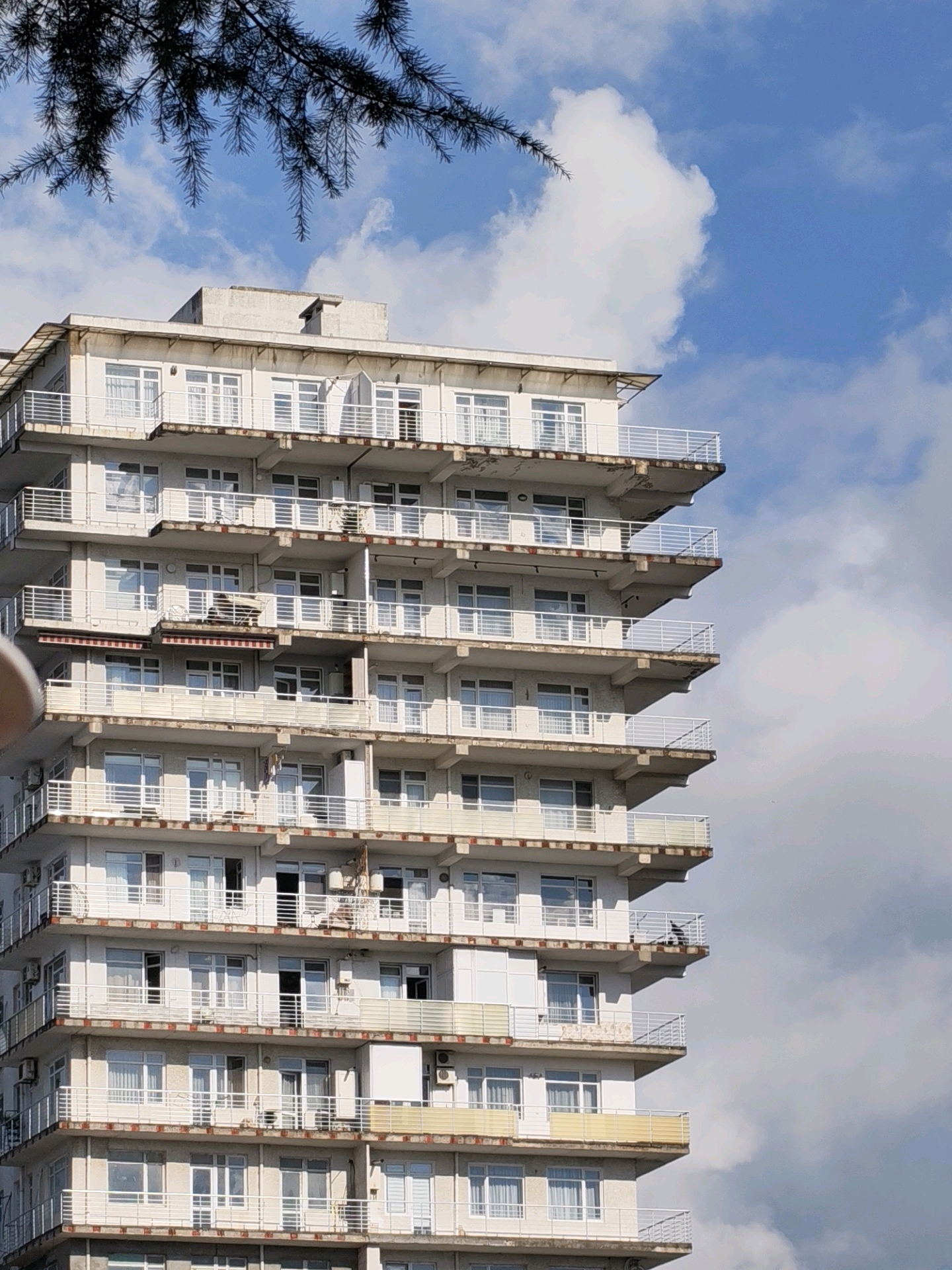 An external, eye-level, medium shot captures a weathered, multi-story building with numerous balconies against a partly cloudy blue sky. The building, likely an apartment complex or hotel in Batumi, Georgia, exhibits signs of age with chipped paint and stained concrete on its balconies and facade. Each floor is lined with white railings, and many balconies are equipped with outdoor furniture and air conditioning units. A few windows are open, revealing curtains and glimpses of the interiors. A dark, leafy tree branch is visible in the upper left corner, partially obscuring the sky. The lighting suggests it is daytime, with shadows cast on the building's surfaces.