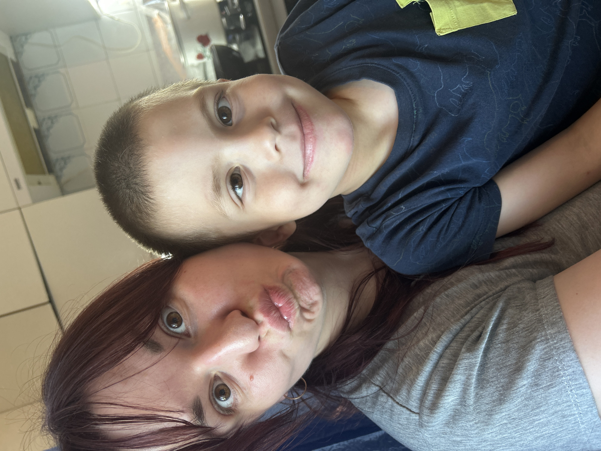 The image shows a close-up selfie of two people, a child and an adult, lying down together. The child, a boy, is positioned above the adult, with his head resting on the adult's shoulder or chest. He is looking directly at the camera with a slight smile. He has short, dark hair and brown eyes. He is wearing a dark blue t-shirt with a yellow emblem on the chest.
The adult, likely a woman, is positioned below the child and is looking at the camera with her lips pursed in a playful expression. She has long, reddish-brown hair that frames her face. She is wearing a grey t-shirt. Her eyes are wide open, and she appears to be making a funny face.
The background is blurry but appears to be an indoor setting, possibly a home. The lighting suggests it could be daytime. There are no obvious external cues to indicate a specific time of day or weather conditions. The overall mood of the image is intimate and playful, suggesting a moment of connection between the child and the adult. The orientation of the image is sideways.
