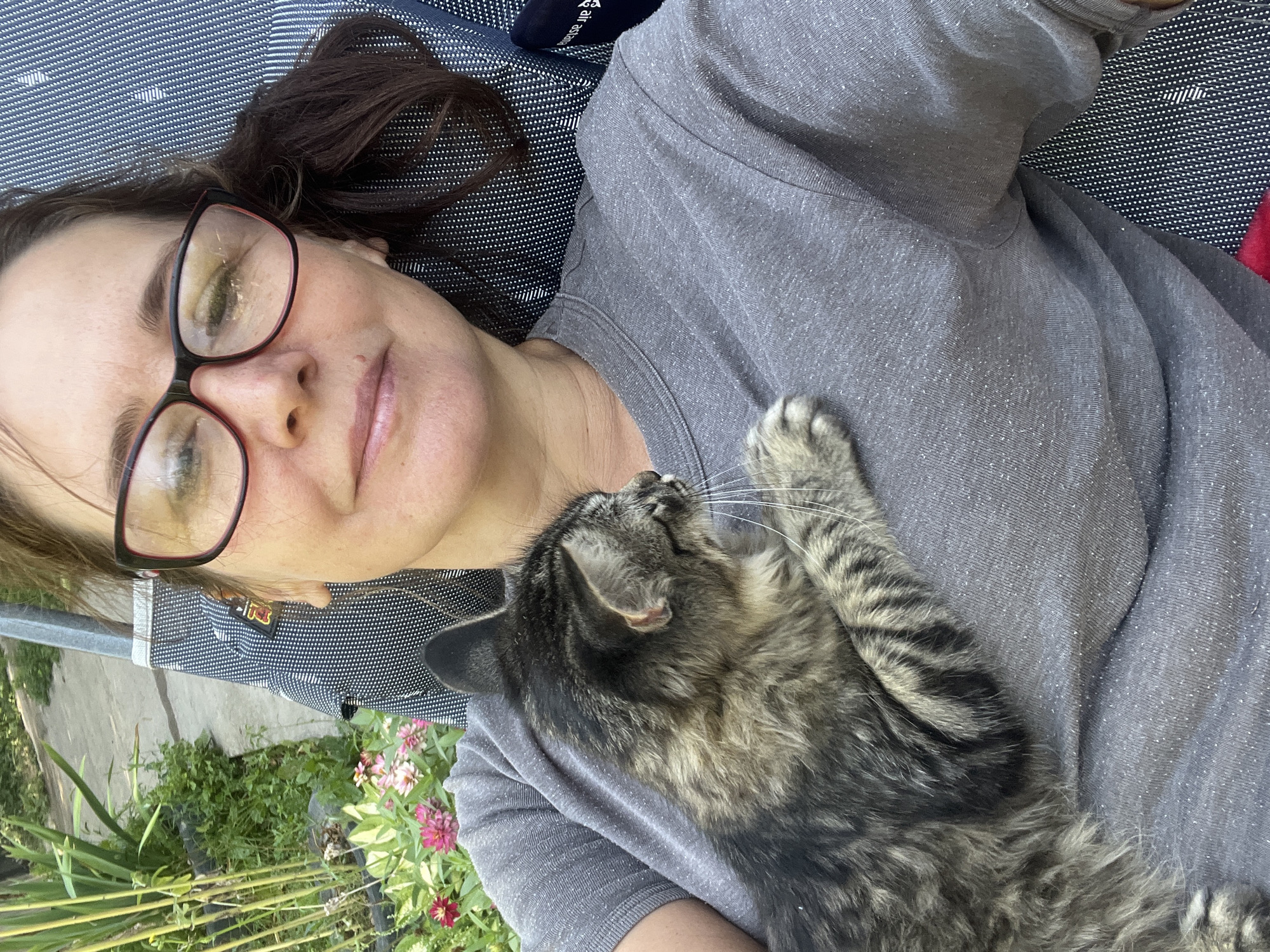 A medium shot, taken from a high angle, shows a woman lying down outdoors, holding a tabby cat. The woman, wearing glasses and a gray t-shirt, is looking up at the camera, smiling. The cat, perched on her chest, is facing to the left and appears to be looking away from the camera. The background suggests a backyard setting, with green plants and flowers visible. The image is captured in Қарғалы, Kazakhstan, with the outdoor setting indicating it could be a residential area. The overall mood is casual and relaxed, with the woman and her cat enjoying a moment together in nature.