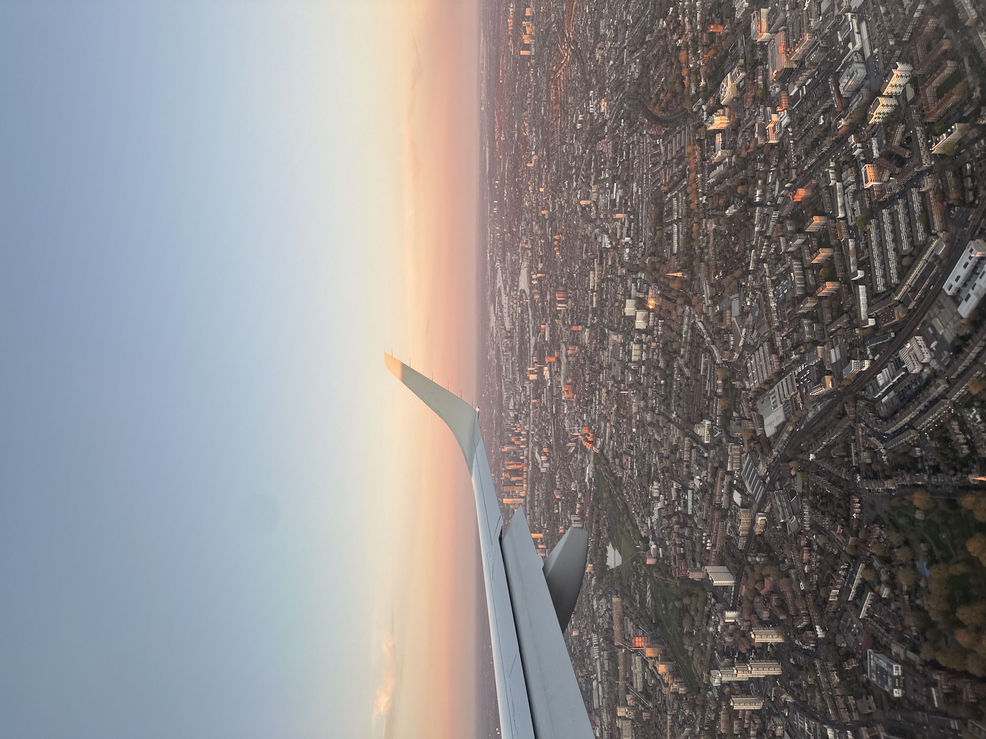 The image shows an aerial view of a densely populated urban area. A portion of an airplane wing is visible in the foreground, suggesting the photo was taken from an aircraft. The city below appears to be London, United Kingdom, given the architectural styles and layout. It seems to be taken over a residential area with many buildings. A park area is visible. The sky is a gradient of colors, suggesting a sunset or sunrise.