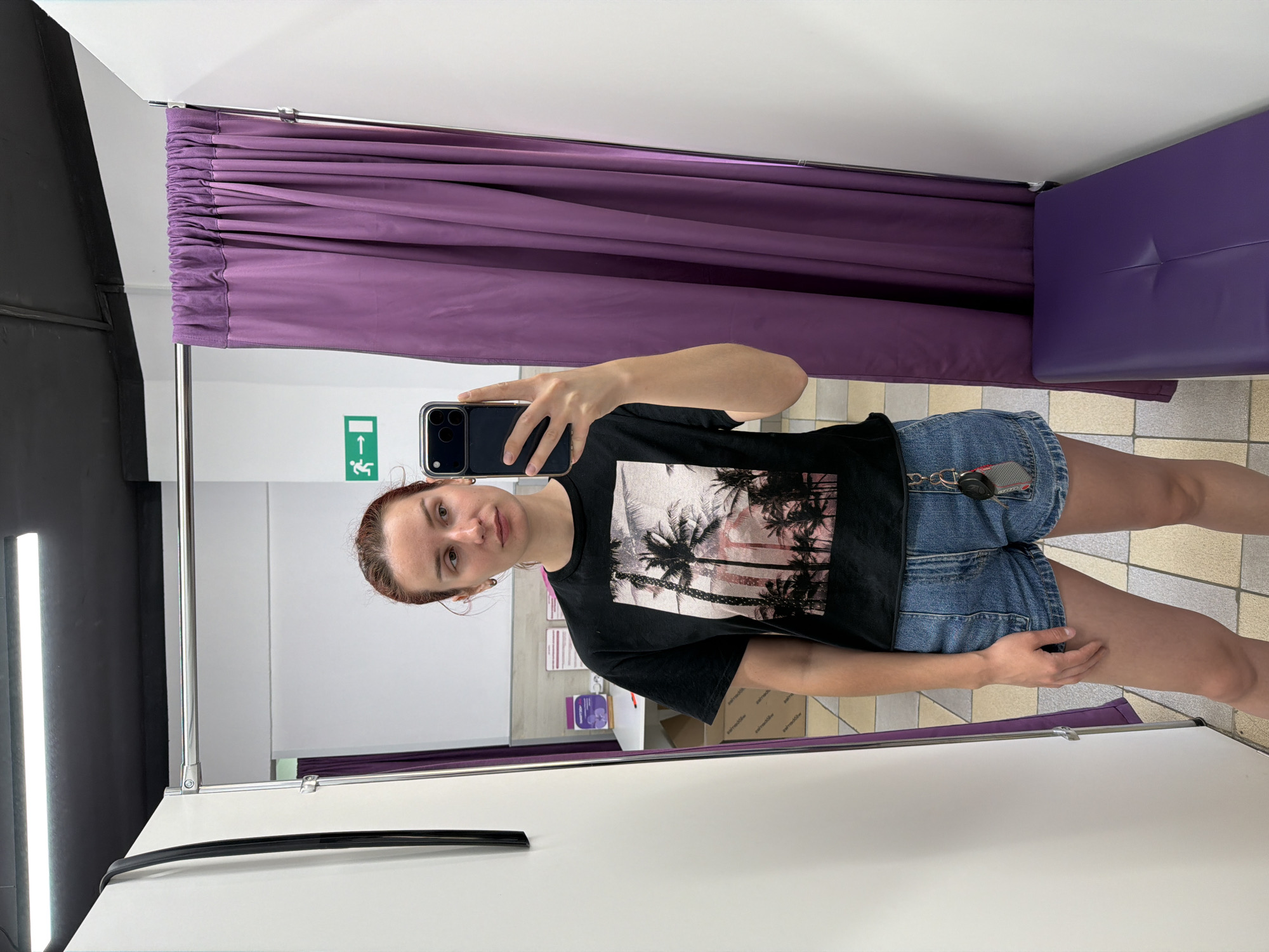 A woman is taking a selfie in a fitting room. She has reddish-brown hair and is wearing a black t-shirt with a palm tree graphic and denim shorts. The fitting room has a purple curtain and a cushioned seat. An emergency exit sign is visible on the wall. Based on online references to stores fitting the description of having purple curtains and seating, the location may be within a Pepela store in the city of Chaikovsky, Russia.
