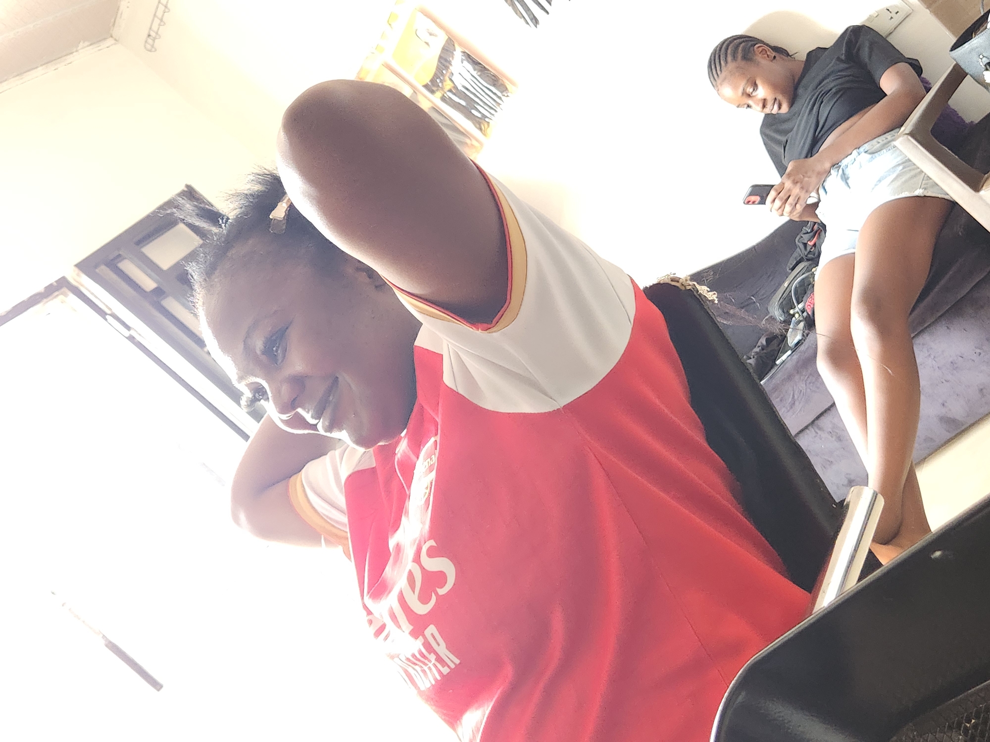In Sabon Tasha, Nigeria, a low-angle shot captures two individuals. The foreground features a person lying back with their hands behind their head, wearing a red shirt with white and yellow trim. White text, possibly a logo or name, is visible on the shirt. To the right and further back, another individual is seated and looking at their phone, wearing a black t-shirt and light-colored denim shorts. The environment appears to be indoors, with bright lighting suggesting daylight. Some items are visible in the background, including what seems to be a package of hair accessories. The overall scene is casual and relaxed.