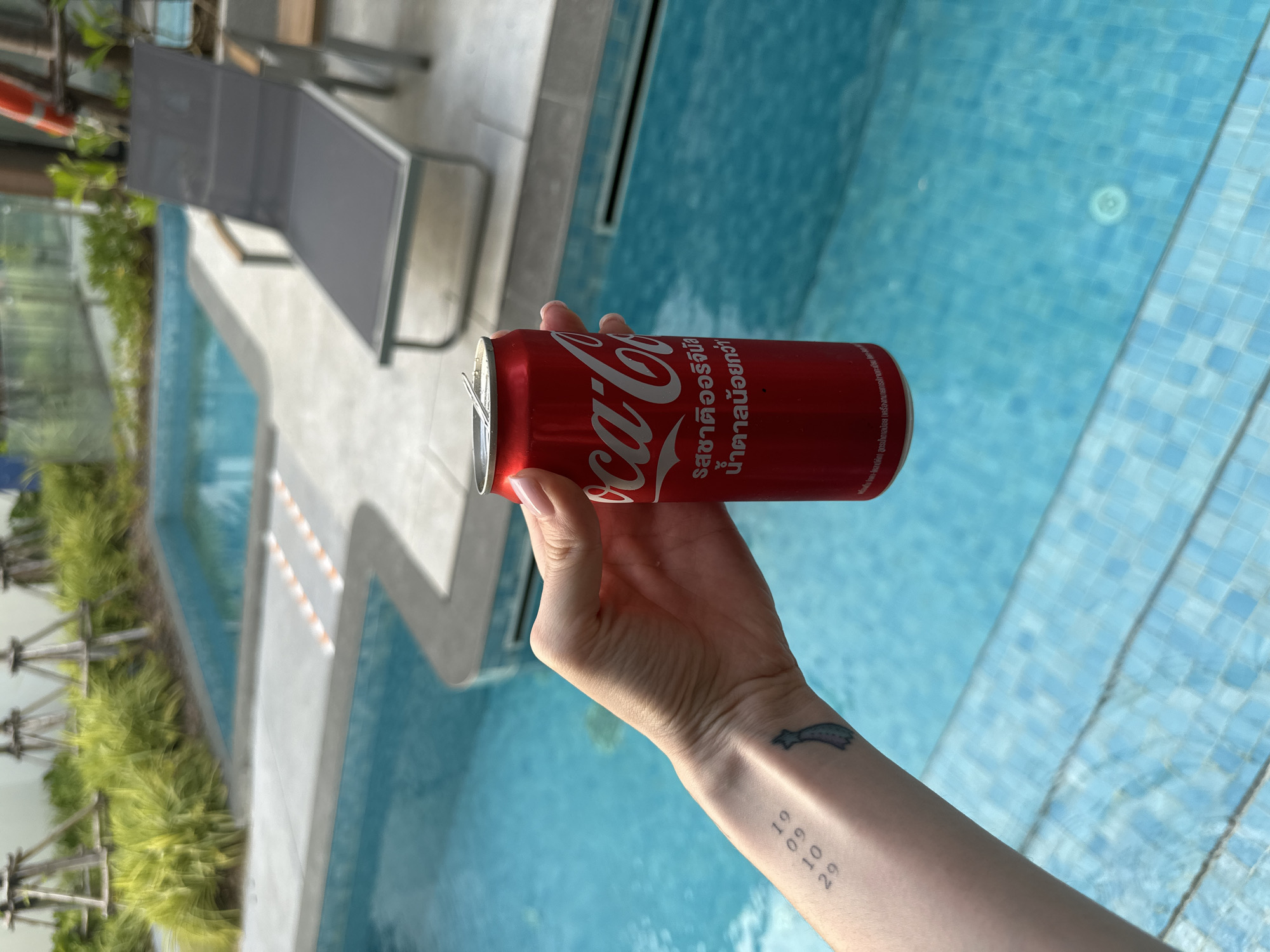 A forensic examination of the image reveals a partial view of a person's right arm and hand holding an open red Coca-Cola can. The can displays the Coca-Cola logo in white and Thai script reading "Original taste, less sugar." The hand exhibits neatly manicured fingernails. Two tattoos are visible on the forearm: one showing the numbers "19 09 29" vertically stacked, and another depicting a stylized blue/grey object resembling a tie or ribbon.
The immediate environment consists of a light blue tiled swimming pool with clear water, bordered by light grey concrete or tiled paving. In the background, a dark grey mesh lounge chair with wooden armrests is positioned poolside. Further back, green foliage, possibly ornamental grasses, lines parts of the pool area, and young trees supported by wooden stakes are visible. The setting suggests an outdoor leisure area, likely at a resort or residence in the city of Choeng Thale, Thailand.