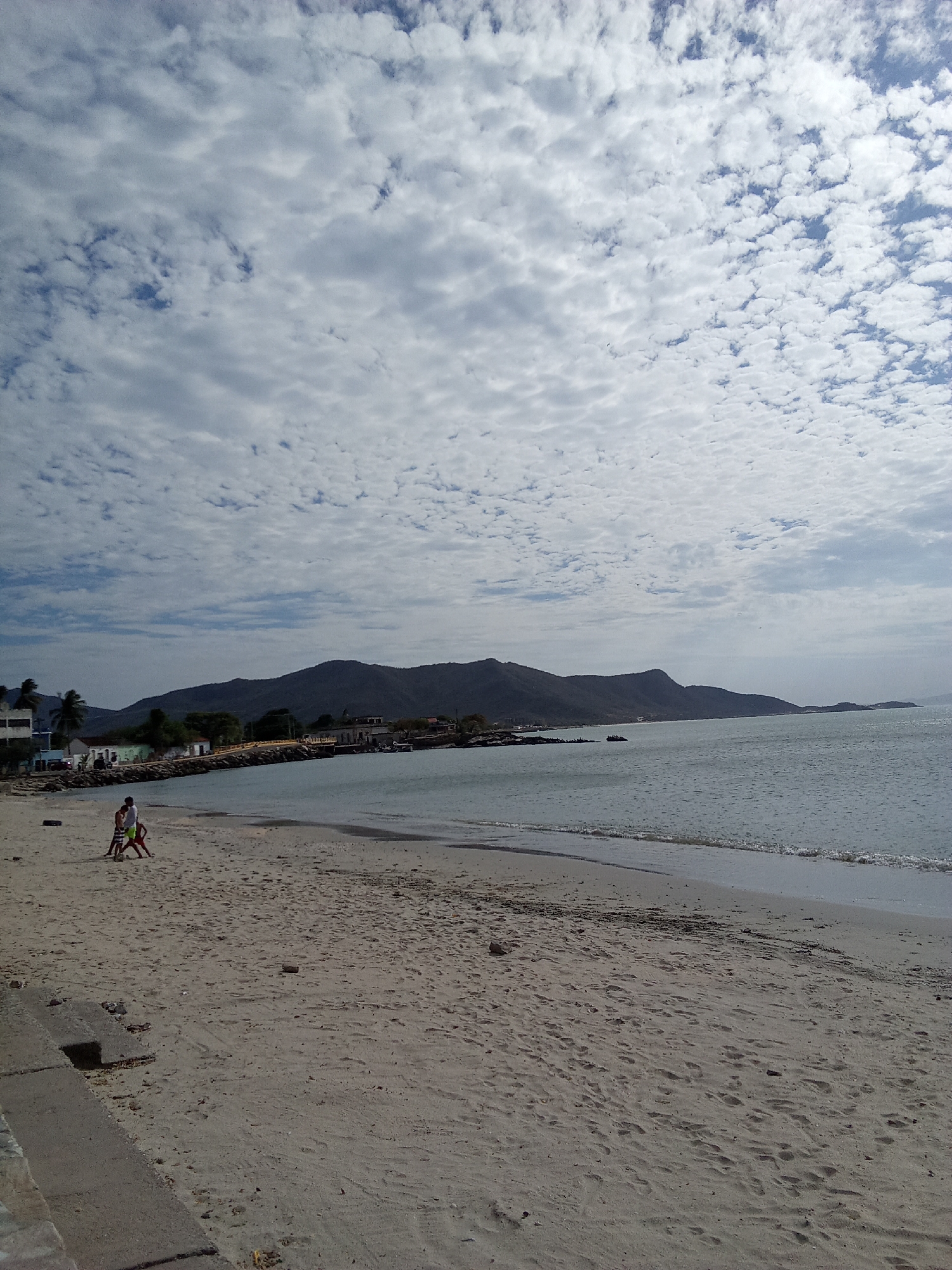 The image depicts a beach scene in Juan Griego, Venezuela. In the foreground is a sandy beach with a concrete wall on the left. Two people, a child and an adult, are standing near the shoreline. The calm water of the bay stretches towards the horizon, where a mountainous coastline is visible. The sky is filled with scattered clouds.