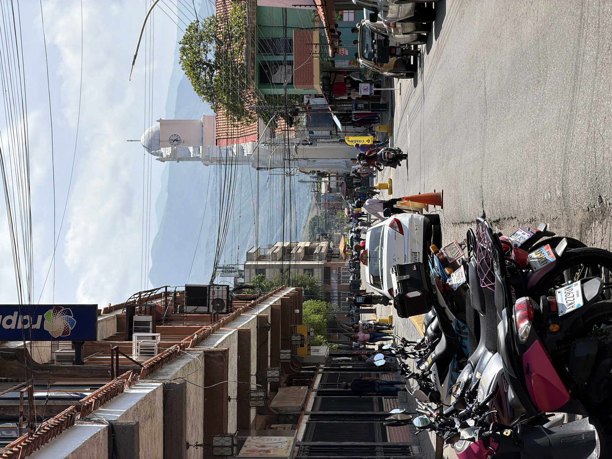 In Turmero, Venezuela, a street scene unfolds under a bright, partly cloudy sky, with mountains visible in the distance. The street is lined with buildings, some featuring awnings and shops. One building has a blue sign with a colorful logo and the word "rciper". Air conditioning units are visible on the exterior of some buildings. A white sedan with a yellow "TAXI" sign on its roof is parked on the right side of the street. Several motorcycles and scooters are parked along the left side of the street. Pedestrians are walking on the sidewalk and in the street. A clock tower, part of a white building with a red roof, stands out in the background. The street is paved and shows signs of wear. There are utility wires overhead, characteristic of urban infrastructure. The overall impression is of a typical day in a Venezuelan city, with a blend of commerce, transportation, and everyday life.