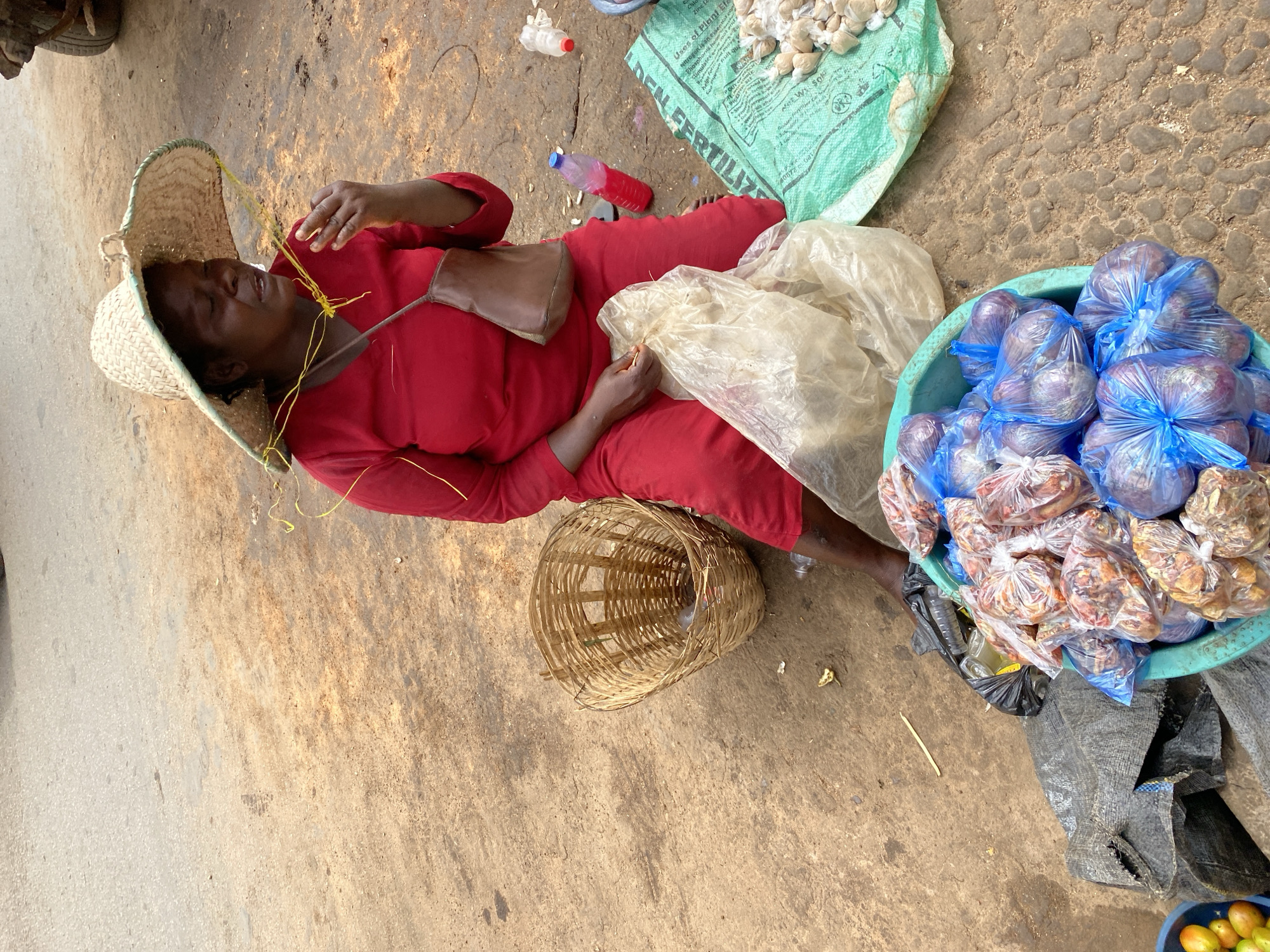 In Bukuru, Nigeria, a woman wearing a wide-brimmed straw hat and a red outfit sits on the ground beside a collection of goods. She is holding a small item to her mouth and has a brown satchel slung over her shoulder. In front of her, a turquoise basin is filled with onions wrapped in blue plastic bags. To the right, a green bag labeled "DEN FERTILIZER" lies open, displaying a pile of small white items, likely garlic or ginger. A woven basket sits to her left. The ground is a mix of asphalt and dirt, with scattered debris.