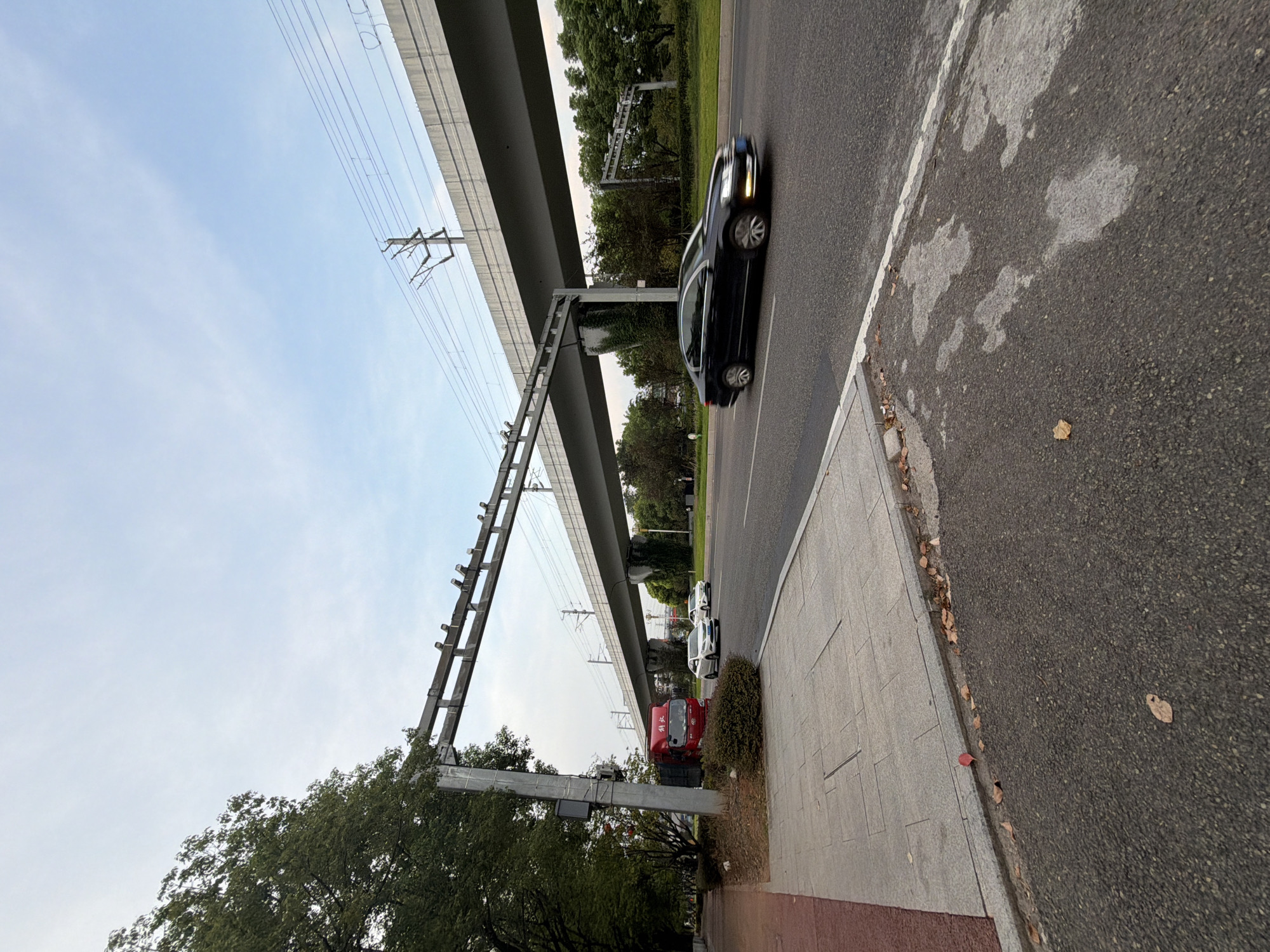 The image depicts an outdoor scene on a cloudy day, featuring a multi-lane road beneath an elevated structure, likely an overpass or train track, in Jiangjin, China. On the left side of the road, a sidewalk with gray paving stones is visible, bordered by a red-colored pedestrian lane. Adjacent to the sidewalk is a lush green tree. A red truck is parked on the side of the road near the sidewalk. Several cars are present on the road; a black sedan is in motion on the right lane, and two white sedans are parked or moving slowly on the left lane, further down the road. The elevated structure is supported by concrete pillars, and power lines are visible running above it. The road surface is asphalt with some water stains or patches. Fallen leaves are scattered on the sidewalk and road's edge, suggesting an autumn season. The sky is overcast with a pale blue hue.