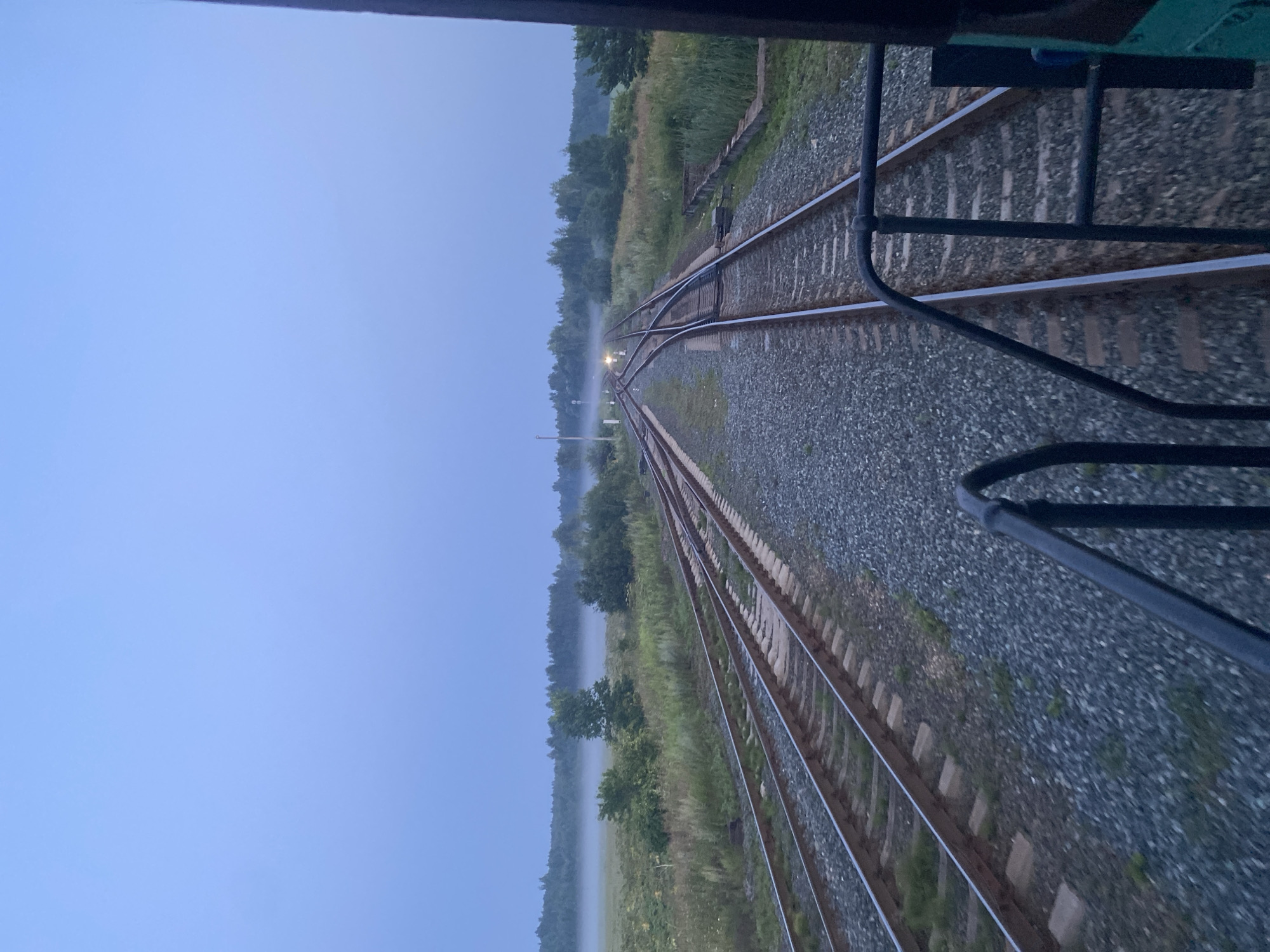 The image depicts a foggy railway landscape at dusk, likely in Kauchuk Stantsiya, Russia. Multiple sets of train tracks converge and diverge, with one set in the foreground showing a weathered appearance and gravel ballast. Beyond the tracks, a grassy field stretches towards a dense line of trees bordering a foggy expanse in the distance. The sky is a pale, hazy blue, indicating twilight or overcast conditions. There are no visible people in the image. The scene is captured from the perspective of someone on a train, with the railing of the train car visible in the foreground. The overall atmosphere is serene and slightly melancholic due to the fog and dim lighting.