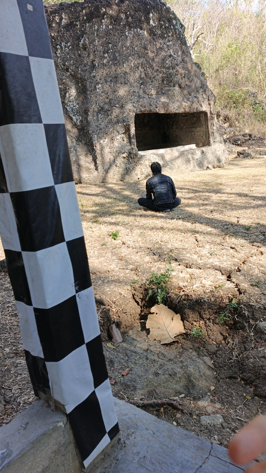 An outdoor, daytime scene depicts a person sitting in front of a large rock formation with a rectangular opening. The rock is textured and weathered, with some vegetation growing on its surface and around its base. The ground is dry and covered with sparse grass and leaves. In the foreground, a black and white checkered pole is visible, leaning against a concrete structure. The person, viewed from behind, is wearing a dark denim jacket and black pants, sitting cross-legged on the ground. The overall environment suggests a historical or archaeological site. The location is identified as Junjung, Indonesia.