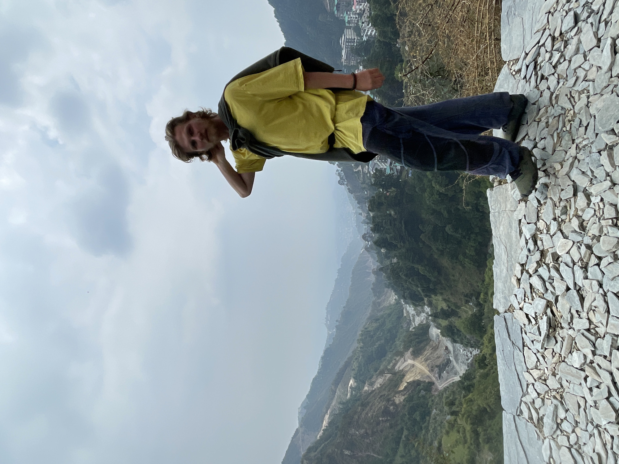 A medium shot captures a man standing on a rock structure in Bhagsu, India. He wears a yellow t-shirt, dark pants, and green shoes with a dark jacket slung over his shoulder. The man is leaning slightly, with his head tilted and his hand near his ear. The backdrop showcases a sprawling mountain range under a cloudy sky. The foreground features a wall constructed of grey rocks. The environment includes lush green foliage and structures typical of Bhagsu.