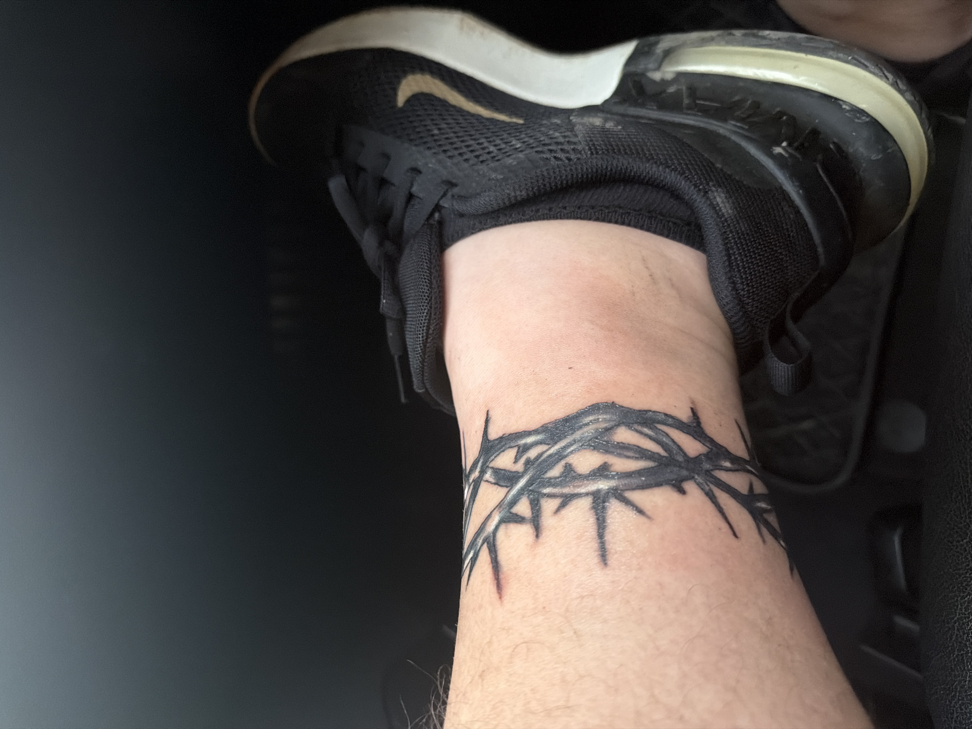 A close-up shot captures a person's lower leg and foot, primarily focusing on a distinctive tattoo and footwear. A prominent black and grey tattoo of a crown of thorns encircles the ankle/lower shin, featuring an intricate design of thorny branches. The foot is adorned with a black athletic shoe, which appears to be a Nike sneaker, identifiable by the golden/tan "swoosh" logo on its side. The shoe features a mesh upper and a white/cream-colored sole, showing signs of wear or dust. The background is dark and out of focus, suggesting an interior setting with dark, possibly upholstered or carpeted surfaces, reminiscent of a car interior. The lighting is moderate, highlighting the details of the leg, tattoo, and shoe. The person's foot is in a static, resting position. No specific activity or emotions are discernible. The only visible text is the Nike swoosh logo. The image is set in Shumen, Bulgaria.