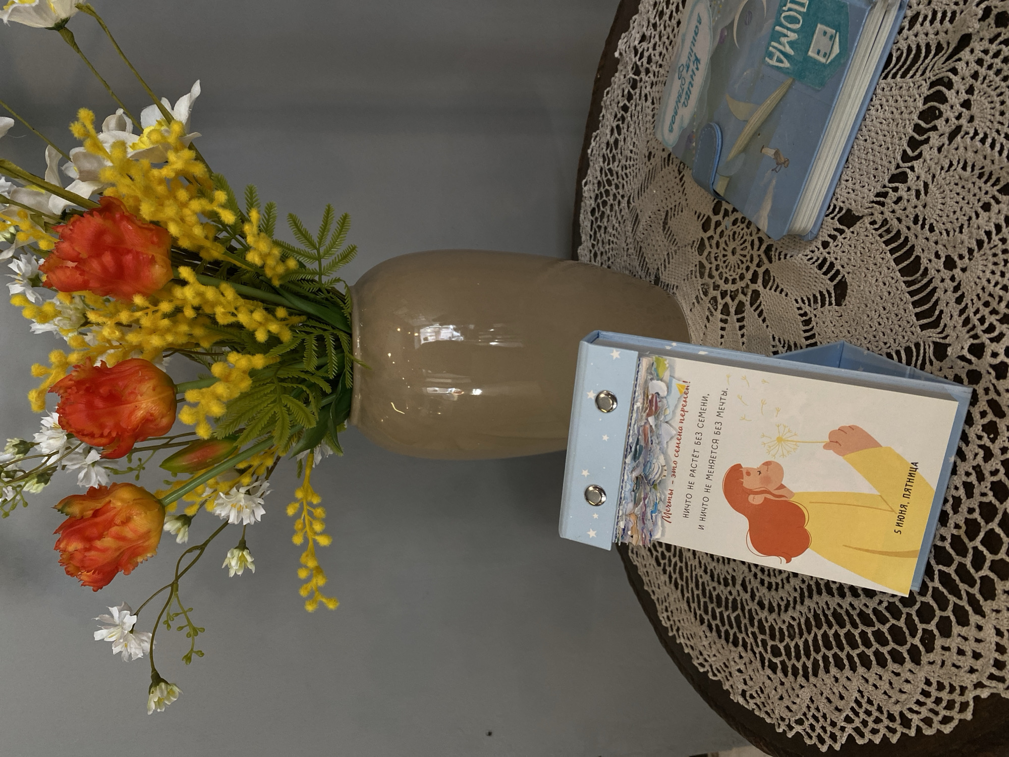 The image presents a still life arrangement on a table with a dark surface, partially covered by a light-colored, intricately crocheted doily. A glossy, light brown vase holds a vibrant floral arrangement featuring ruffled orange tulips, bright yellow mimosa-like flowers, small white blossoms, and green fern-like foliage.
Two light blue books or journals are visible on the doily. One, positioned further back, has "Книга Ваших отзывов" (Book of Your Reviews) and "ДОМА" (Home) visible on its cover. The other, closer to the foreground, is a ring-bound planner or journal. Its cover displays an illustration of a person with reddish-brown hair, wearing a yellow top, blowing a dandelion. Text on this item reads "5 ИЮНЯ, ПЯТНИЦА" (June 5, Friday), followed by the inspirational quote: "Мечты - это семена перемен! НИЧТО НЕ РАСТЁТ БЕЗ СЕМЕНИ, И НИЧТО НЕ МЕНЯЕТСЯ БЕЗ МЕЧТЫ." (Dreams are the seeds of change! NOTHING GROWS WITHOUT A SEED, AND NOTHING CHANGES WITHOUT A DREAM.). The background is a plain gray wall. The image is rotated, showing the objects in a sideways orientation.