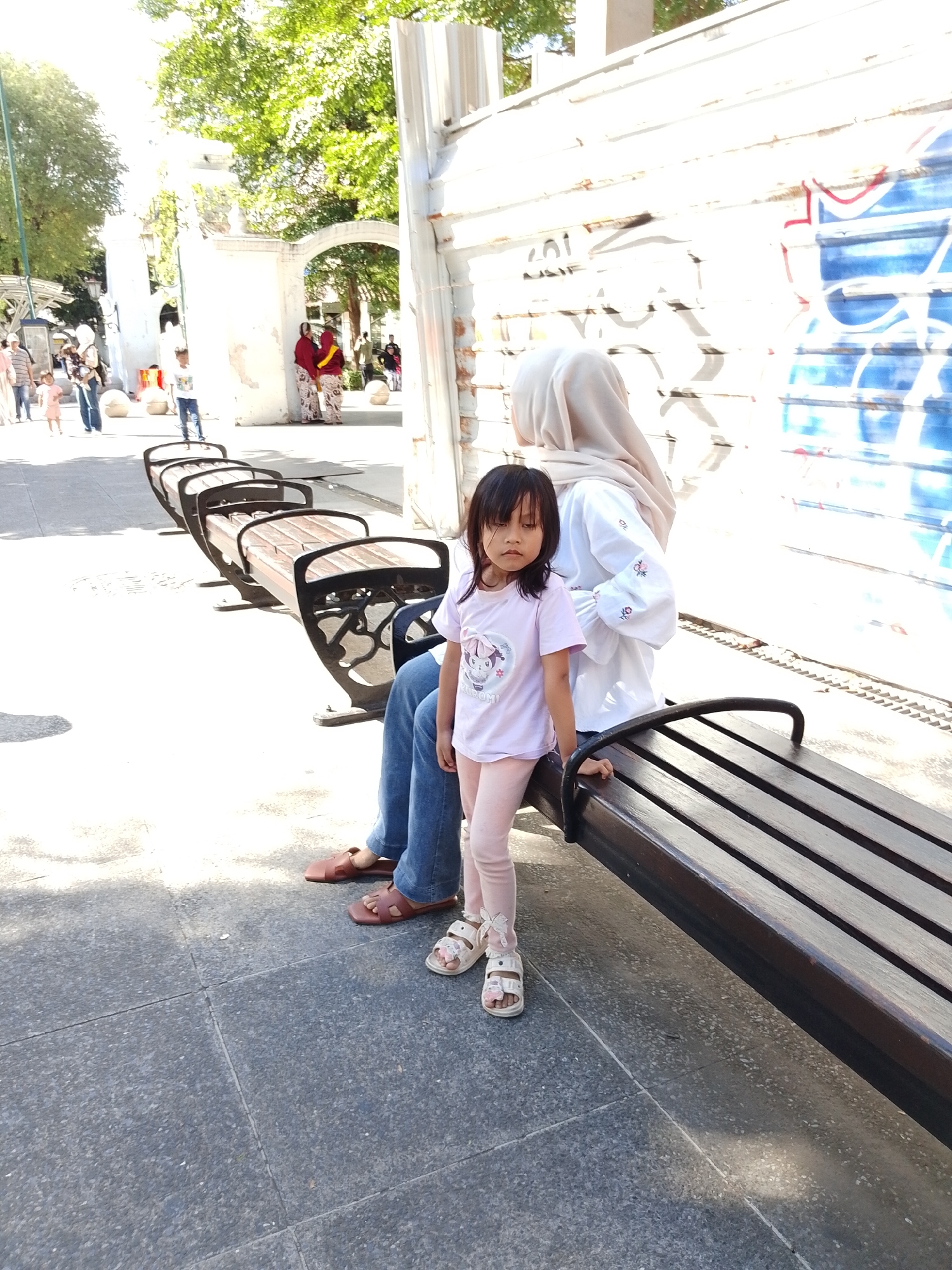 In Yogyakarta, Indonesia, a young girl stands on a paved walkway in front of a row of wooden benches. She is wearing a light pink t-shirt with a cartoon graphic and pink leggings, along with white sandals. Beside her, a person wearing a light-colored hijab and a white shirt with embroidered details sits on the bench, their legs clad in blue jeans. Their feet are shod in brown sandals.
The scene appears to be outdoors on a sunny day, indicated by the bright sunlight and shadows cast on the ground. In the background, there are white archways and buildings, with trees visible overhead. Further in the distance, other people are walking around, some dressed in traditional Indonesian attire. A wall covered in blue and white graffiti is visible to the right. The overall atmosphere seems relaxed and casual, possibly a public park or a pedestrian area in the city.