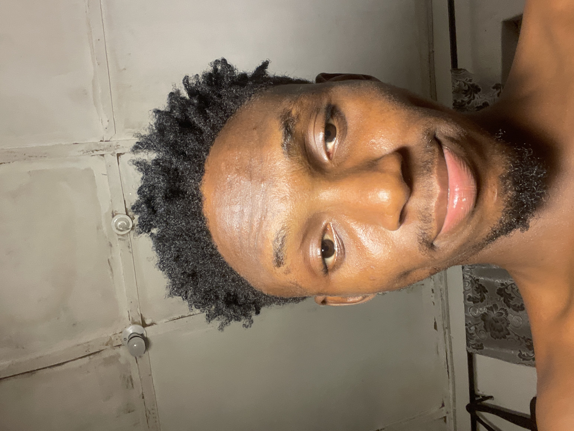 A close-up portrait shows a Black male with short, dark, curly hair and facial hair, including a mustache and beard. His skin appears to have a natural sheen. He is smiling slightly, with his head tilted to the side. The background features a wall with a rough, aged texture and what appear to be ceiling lights. A patterned curtain or backdrop is partially visible. The location, Lokoja, Nigeria is not discernible from this image.