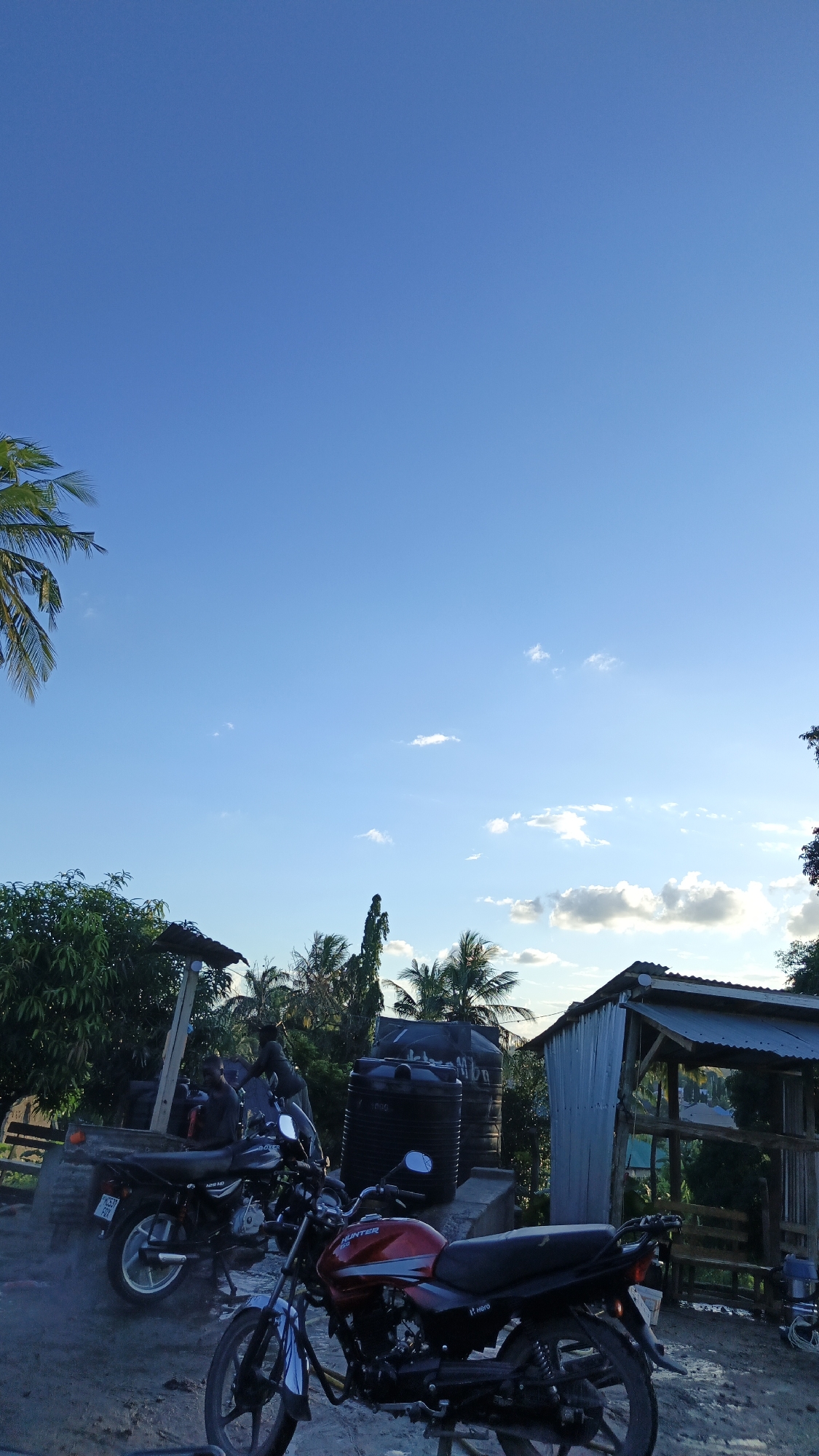 The image presents a scene in an open area in Dar es Salaam, Tanzania, featuring two motorcycles in the foreground. The motorcycle closer to the viewer is red, while the other appears to be black. In the background, two people are near a corrugated iron structure. Two large black water tanks are also visible in the background, alongside lush vegetation and palm trees. The sky is clear blue with scattered clouds.