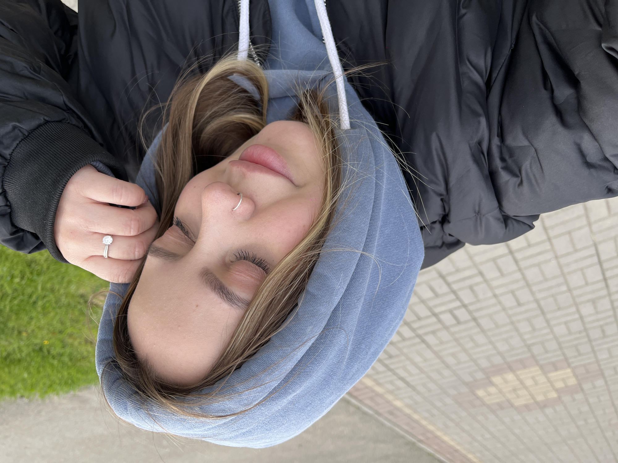 A young woman with closed eyes is lying on a gray paved surface. She is wearing a blue hooded sweatshirt and a black puffy jacket. Her left hand, adorned with a ring, rests near her face. A section of green grass is visible in the upper left corner of the image. The location is Novopokrovskaya, Russia.