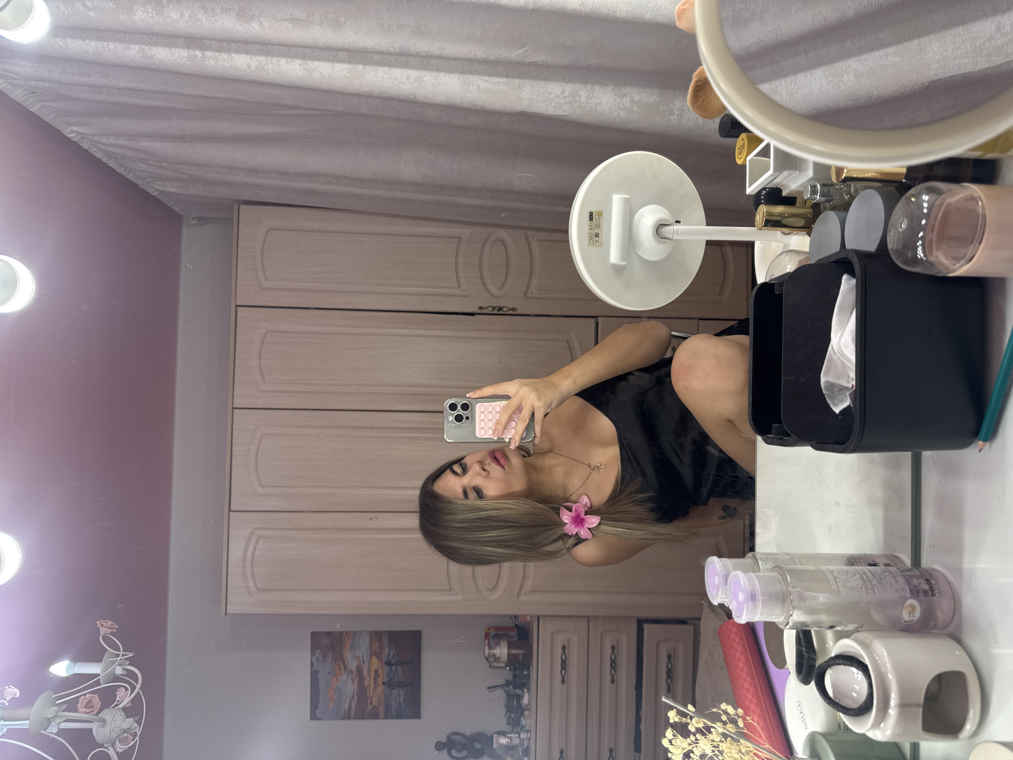 An indoor scene in Chaikovsky, Russia, depicts a young adult female taking a mirror selfie. The woman, with long brown hair highlighted with blonde streaks, wears a black satin camisole and a delicate gold-colored necklace. A pink artificial flower is clipped into her hair on the right side. She holds a light gray iPhone, fitted with a pink checkered case, in her right hand, angled towards her reflection. Her gaze is directed slightly past the phone.
The foreground consists of a white vanity surface holding numerous cosmetic and skincare items. These include multiple clear plastic bottles with light purple caps, one marked "COLOR MILK," a white ceramic holder containing black hair ties, a small bundle of dried flora, a rolled red mat, a black rectangular organizer filled with white tissues or pads, and a back view of a white standing makeup mirror. Various other perfume bottles, cosmetic containers, and makeup brushes are also present, along with a teal pencil and a pink cylindrical container.
Reflected in the mirror, the background reveals a room with a pink ceiling featuring two recessed spotlights and a white chandelier adorned with pink floral accents. Light-colored, textured curtains hang on the right side of the frame. Centrally positioned behind the woman is a light wood, three-door wardrobe with recessed panels. To the left of the wardrobe, a matching chest of drawers with ornate handles supports a framed painting depicting a sunset or sunrise over water with two figures, an abstract dark sculpture, and various small objects. The overall setting is consistent with a domestic bedroom or dressing area.
