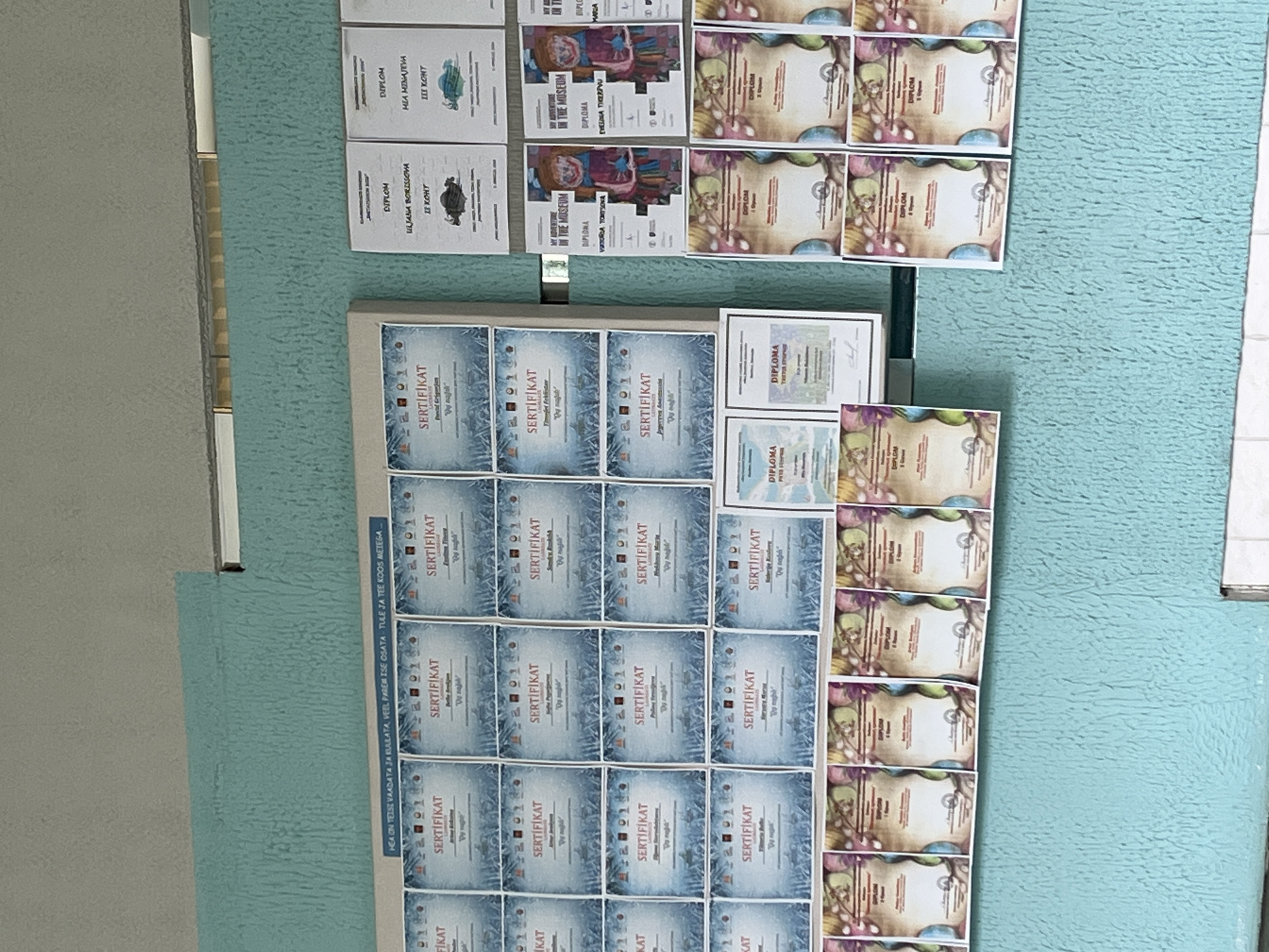 The image shows a wall display in Kohtla-Järve linn, Estonia, featuring multiple certificates and diplomas. The certificates are arranged in rows and columns, predominantly featuring "Sertifikaat" (Certificate) in large lettering. Some certificates have ornate borders and images, while others are simpler. There are also several "Diploma" certificates visible, some of which have colorful decorative elements. The wall itself is painted a textured blue-green color, with a light-colored horizontal band running across the top of the display area. A blue banner with text is visible above the first row of certificates. The overall scene suggests an educational or ceremonial setting where achievements are recognized.