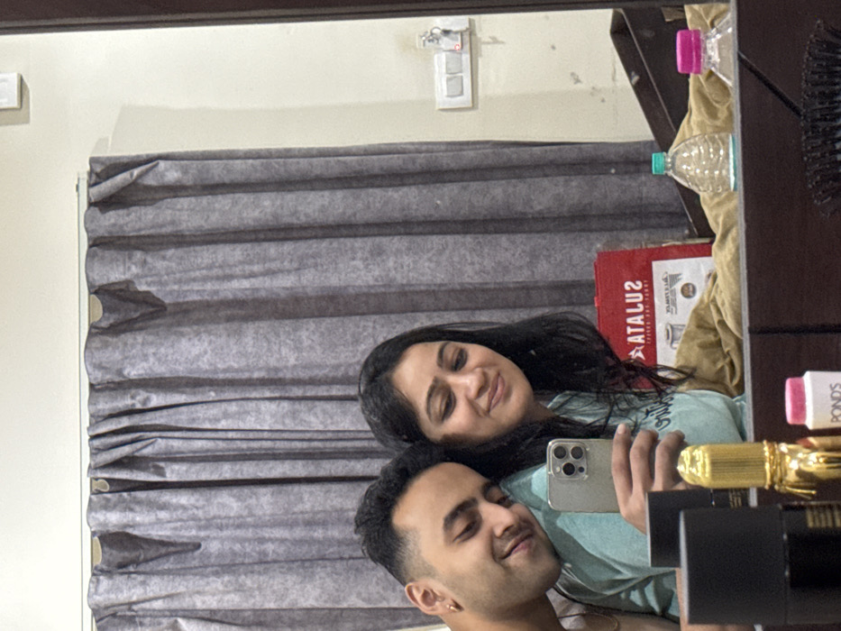 In a room located in Malasar, India, a mirror reflects an intimate scene featuring an adult male and an adult female, both appearing to be of South Asian descent. The female, positioned on the right, holds a silver-colored smartphone with three camera lenses, taking a mirror selfie. She has long, dark hair and wears a light blue top, smiling softly. The male, on the left, rests his head on her shoulder, smiling with his eyes partially closed. He has short, dark hair with a fade and wears a light-colored top. An earring is visible in his left ear.
The background, visible in the mirror's reflection, includes dark grey, textured curtains. To the right of the curtains, a cream-colored wall features a white electrical switch plate with at least two switches, from which a key is suspended. A dark brown vertical frame is present on the far right. On a surface below the reflections of the individuals, various objects are arranged: a black cylindrical item, a gold-colored ornate bottle, a small white container with a pink cap labeled "POND'S", two plastic water bottles (one with a teal cap, one with a pink cap), and a black hairbrush with white bristles. A red shopping bag with reversed white text, "ATALUZ" and "COFFEE AND PERFUMES", rests behind some of these items on what appears to be a light brown or beige fabric.