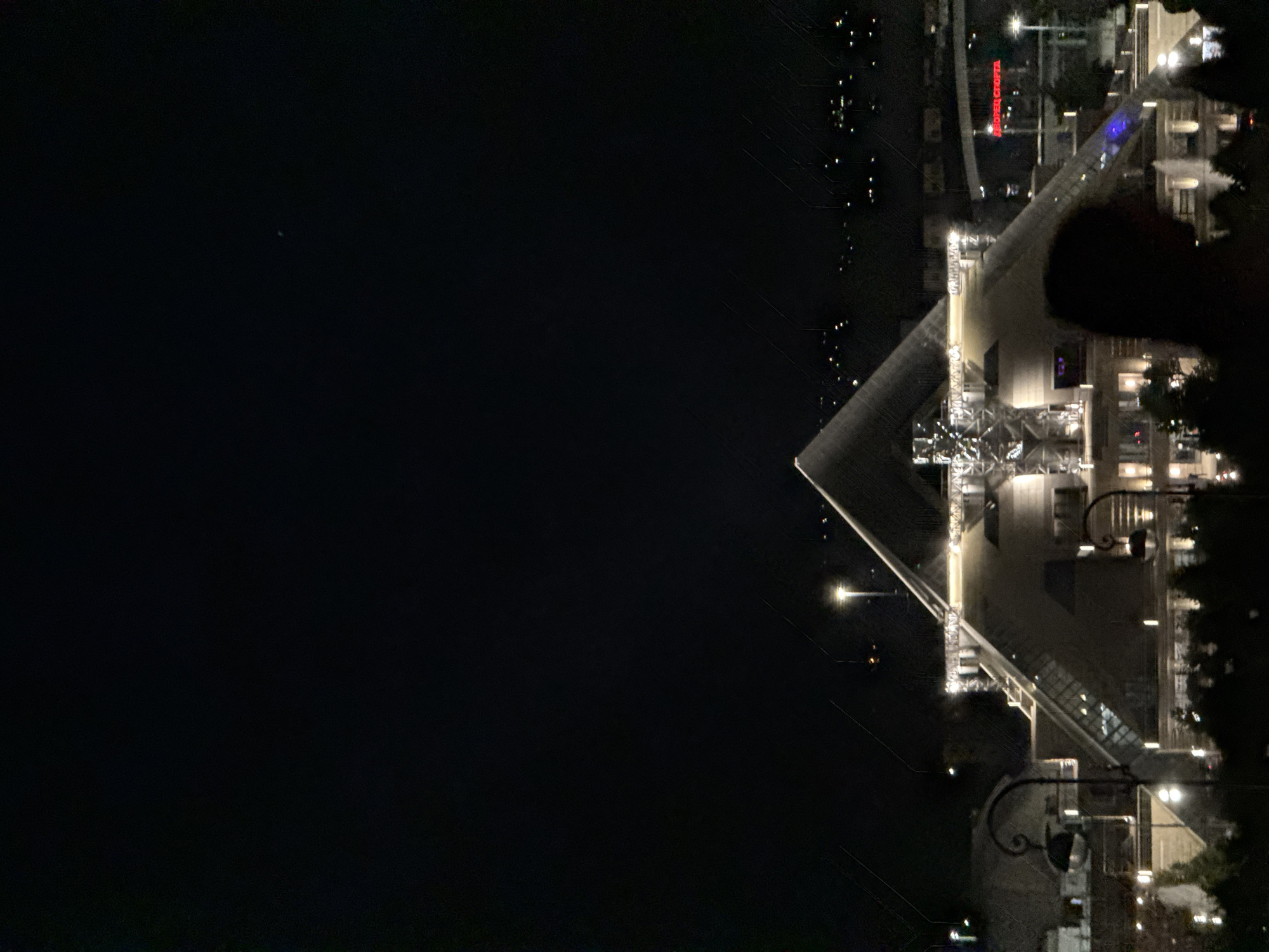 The image shows a nighttime view of Kazan, Russia. Prominently featured is the Kazan Family Center, a large, modern building complex illuminated with bright lights. Adjacent to the Family Center is a body of water, presumably a lake or river, and dark open space, possibly a park or undeveloped land. Small lights can be seen across the water. The image is taken from a high vantage point, providing an overview of the Family Center and surrounding area.