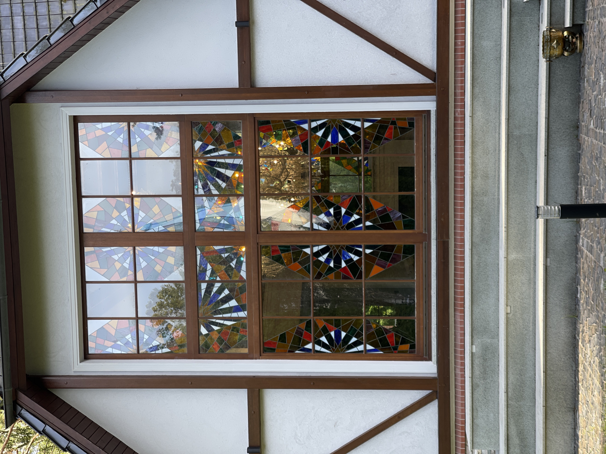 The image shows a large, ornate stained-glass window set into the facade of a building. The window is divided into two sections, each with multiple panes of stained glass featuring geometric patterns in a variety of colors, including red, blue, orange, yellow, and white. The glass is framed by dark brown wood. The building's exterior is white stucco with dark brown trim. Below the window are three wide steps made of grey stone. The steps lead to a paved area constructed with cobblestones. A black cylindrical lamppost stands on the left side of the paved area, and a decorative brass planter sits on the right side. The background shows trees and sky reflected in the stained glass. The scene is an exterior shot of a building in Svetlogorsk, Russia.